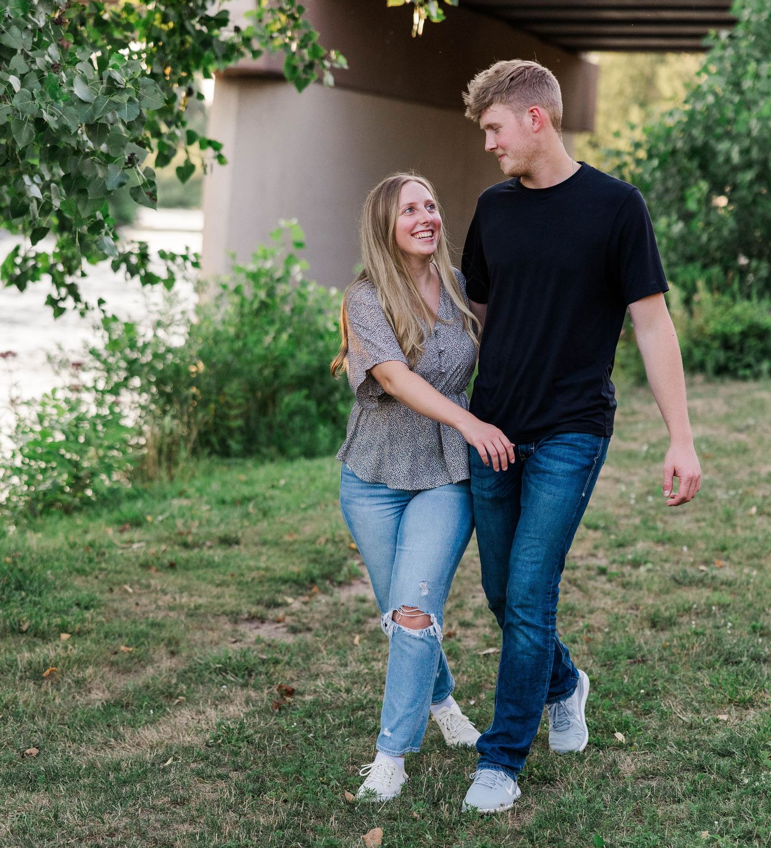 A smiling couple walks hand-in-hand through a lush green park, dressed casually in jeans and t-shirts.
