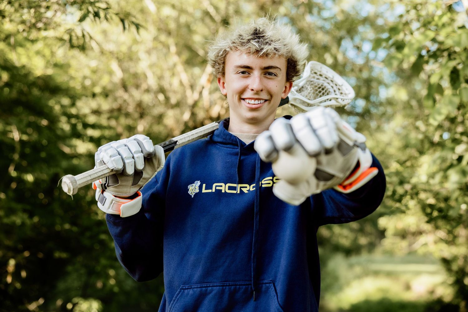 Smiling teen boy in lacrosse hoodie holds stick and gloves outdoors among green trees.