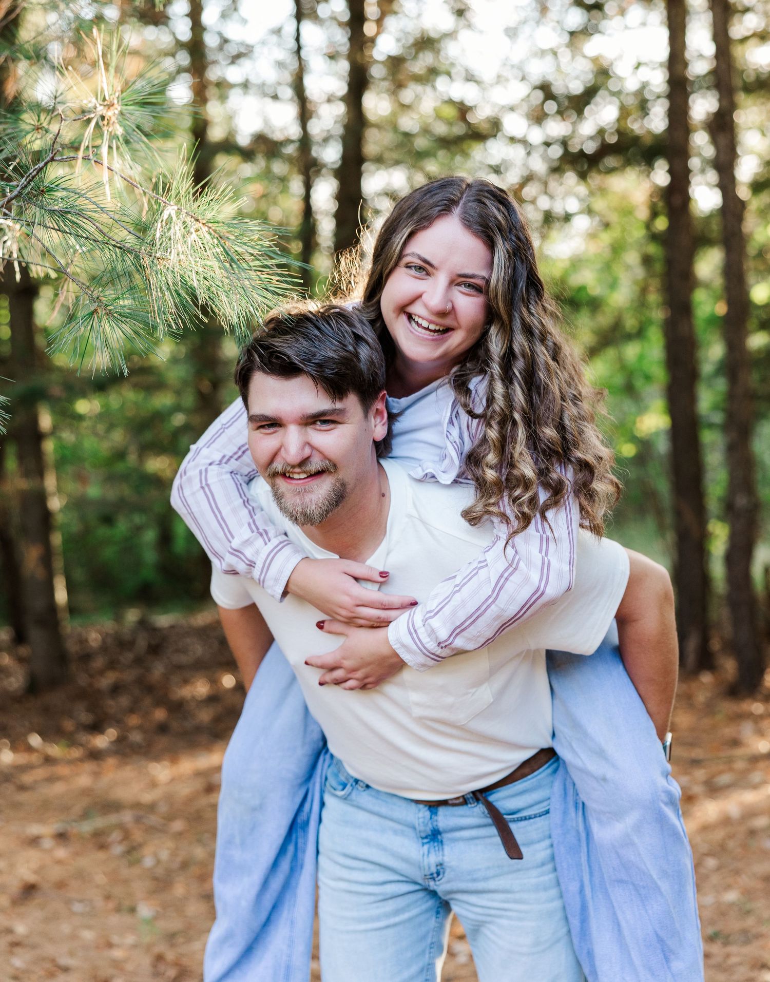 A smiling couple poses in a pine forest, the man giving the woman a piggyback ride.