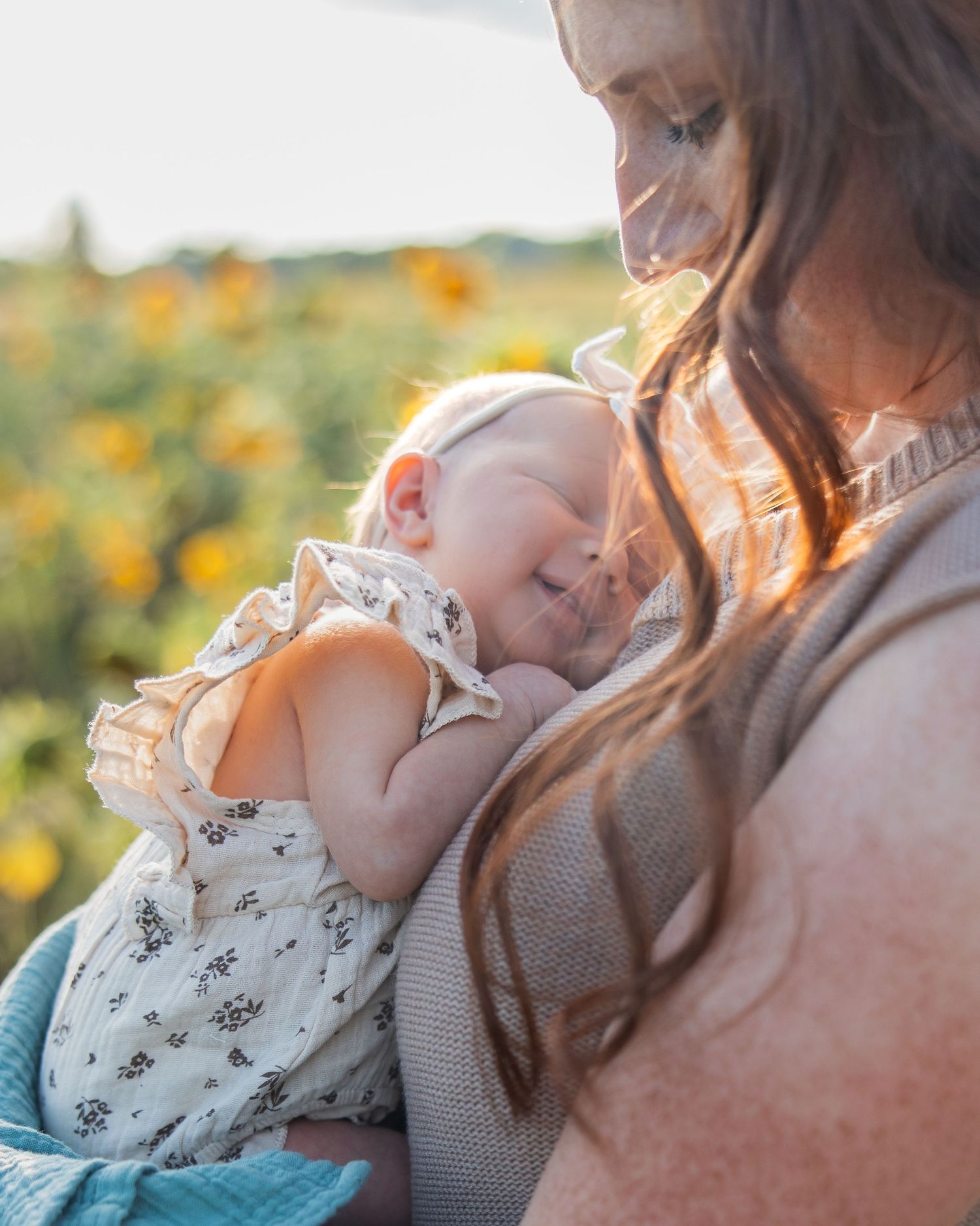 A mother tenderly holds her smiling newborn baby in a sunflower field during golden hour.