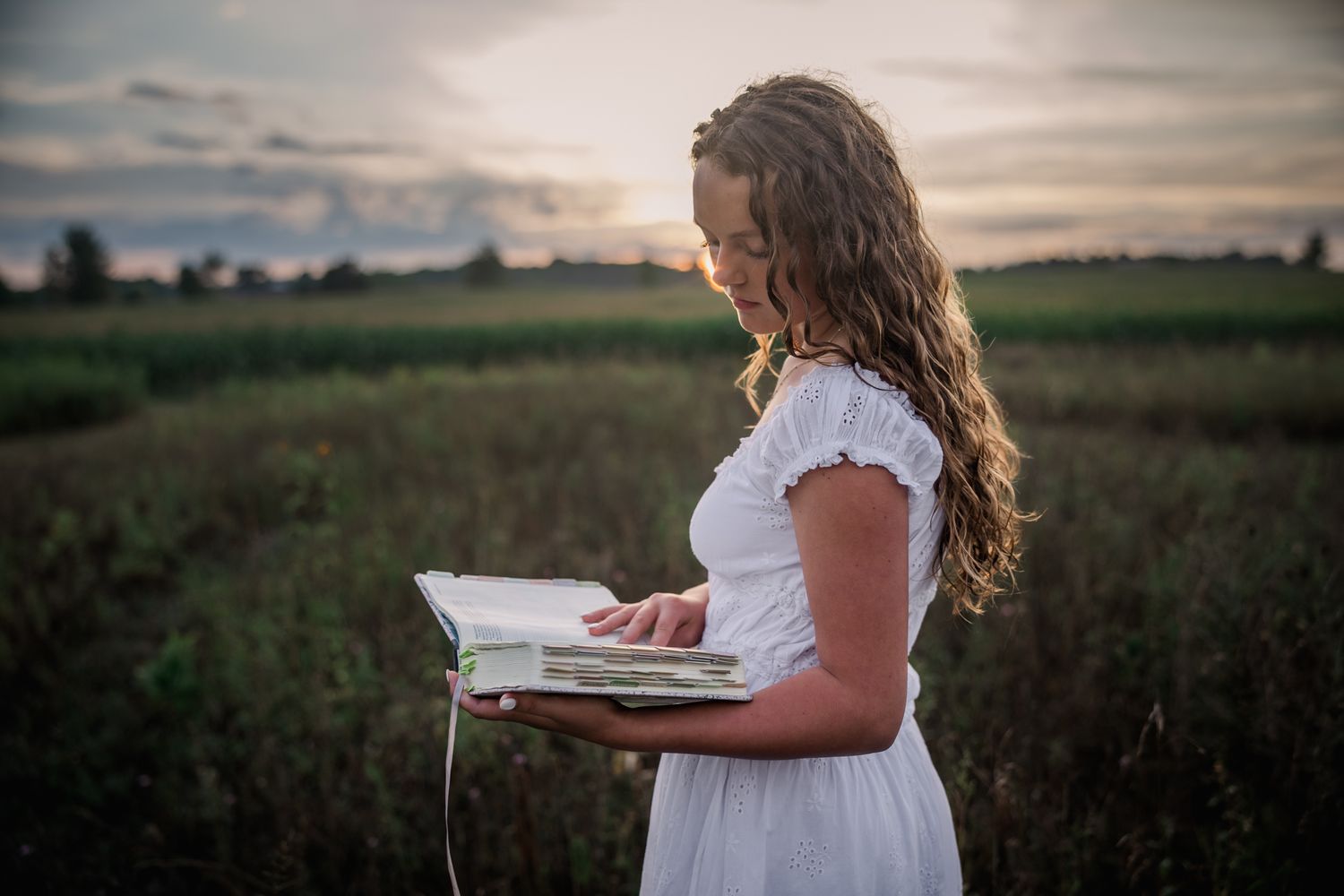 Young woman in white dress reading a book in a green field at sunset.