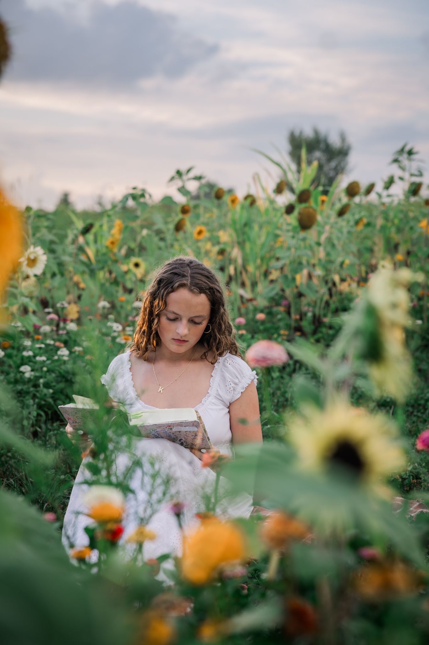 Young girl in white dress reading a book surrounded by colorful sunflowers and wildflowers in a garden at dusk.