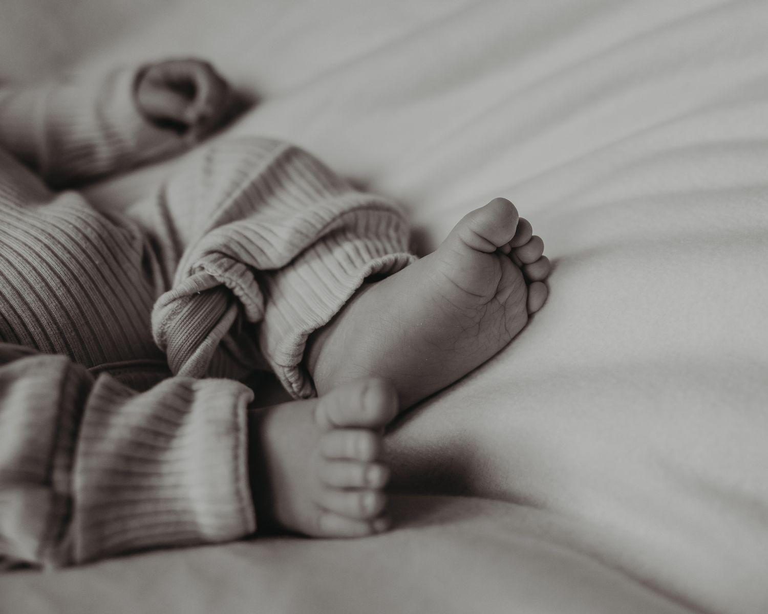Black and white photo of newborn baby feet and hands in soft ribbed onesie on white bedding.