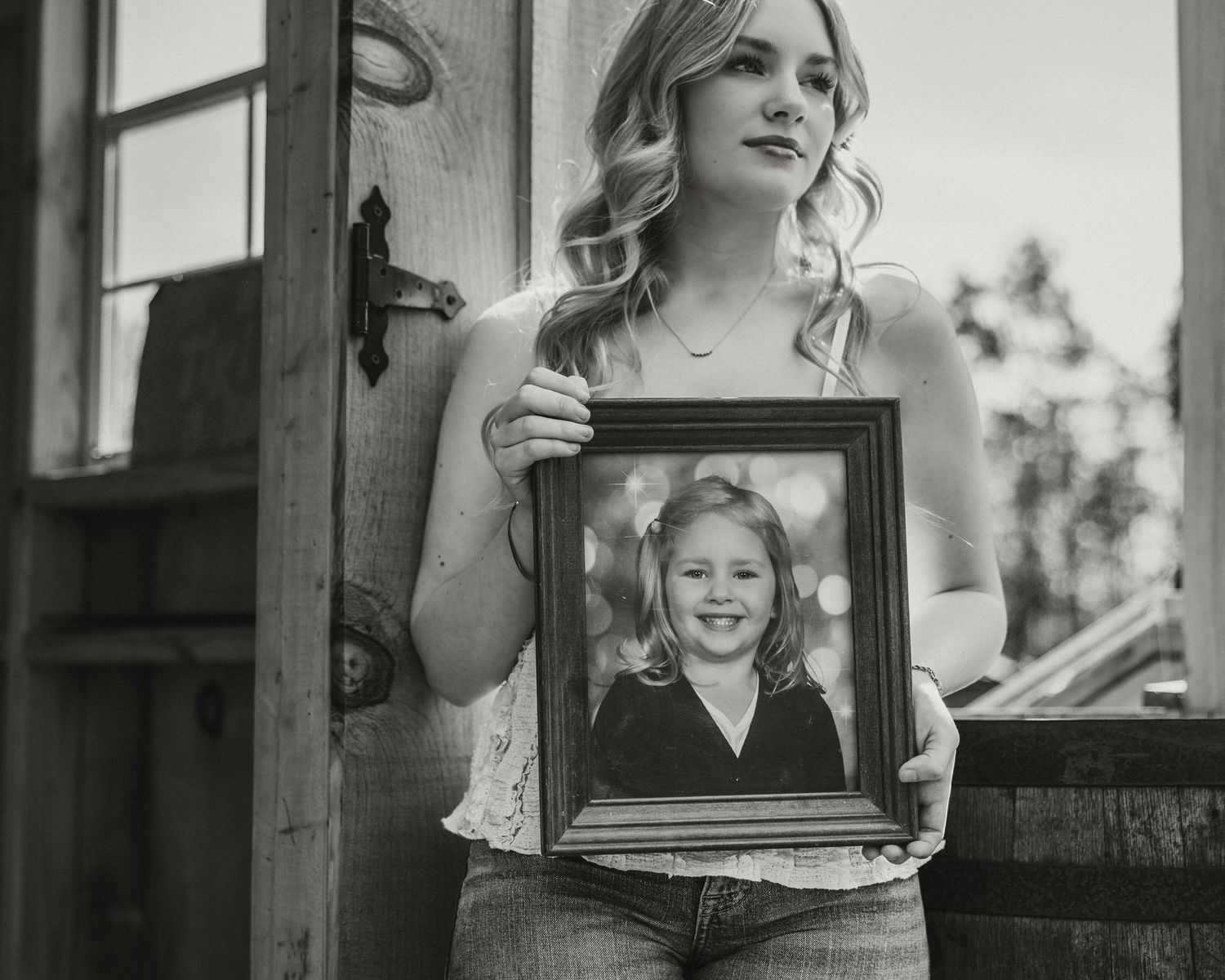 Young woman holding a framed childhood photo of herself in a black and white portrait near a rustic wooden door.