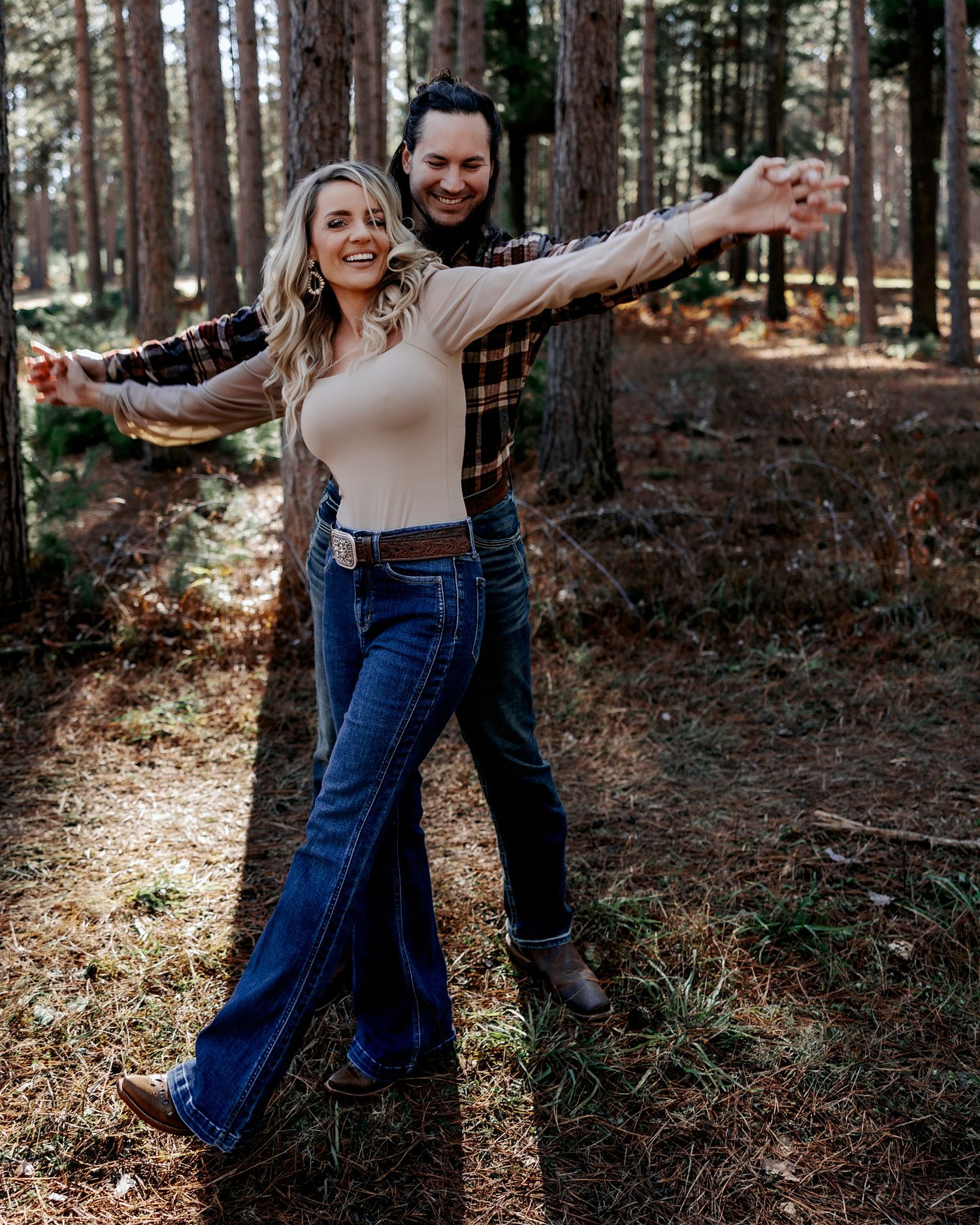Couple joyfully posing in a pine forest, arms outstretched, wearing jeans and plaid shirts.