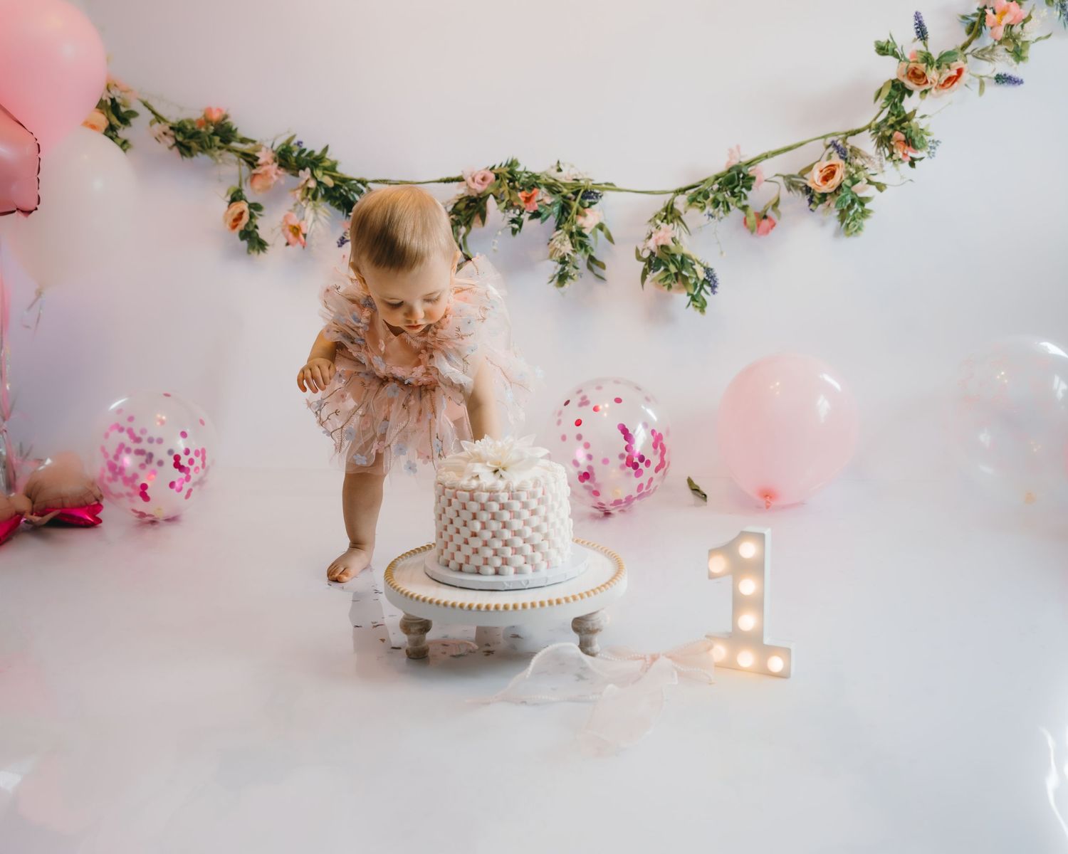 Toddler girl in pink tutu dress stands over white cake with floral garland backdrop at first birthday party.
