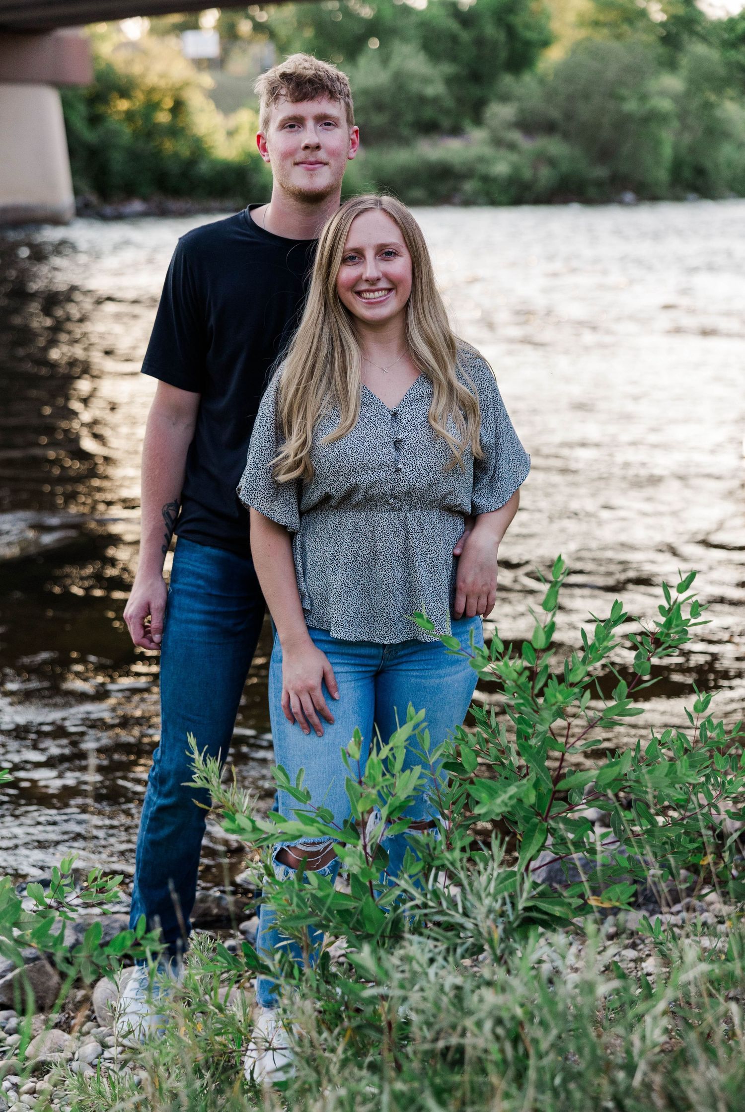 A smiling couple poses together near a river with lush greenery and a bridge in the background on a sunny day.
