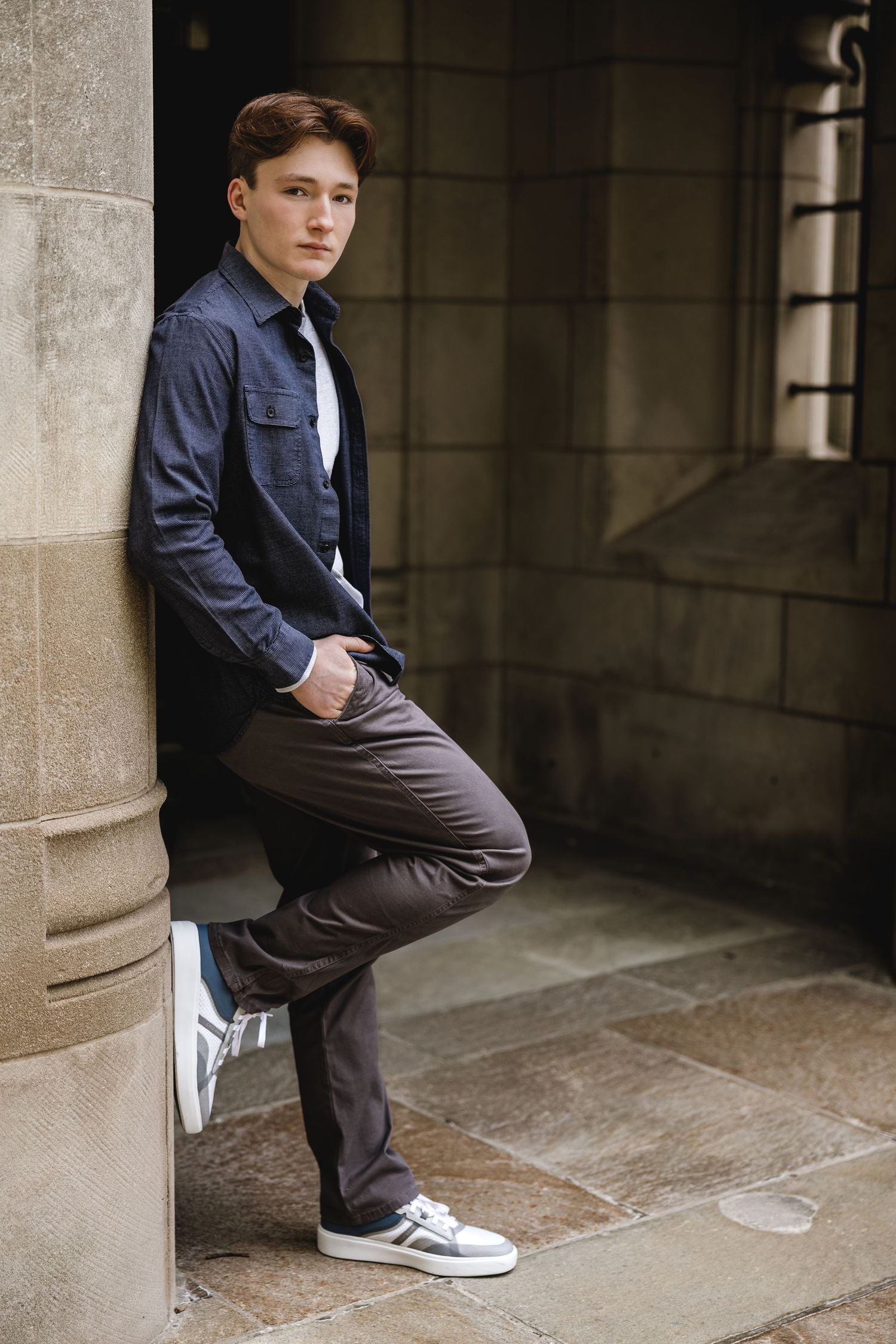 Young man in dark jacket and gray pants leaning against stone wall, posing confidently outdoors.
