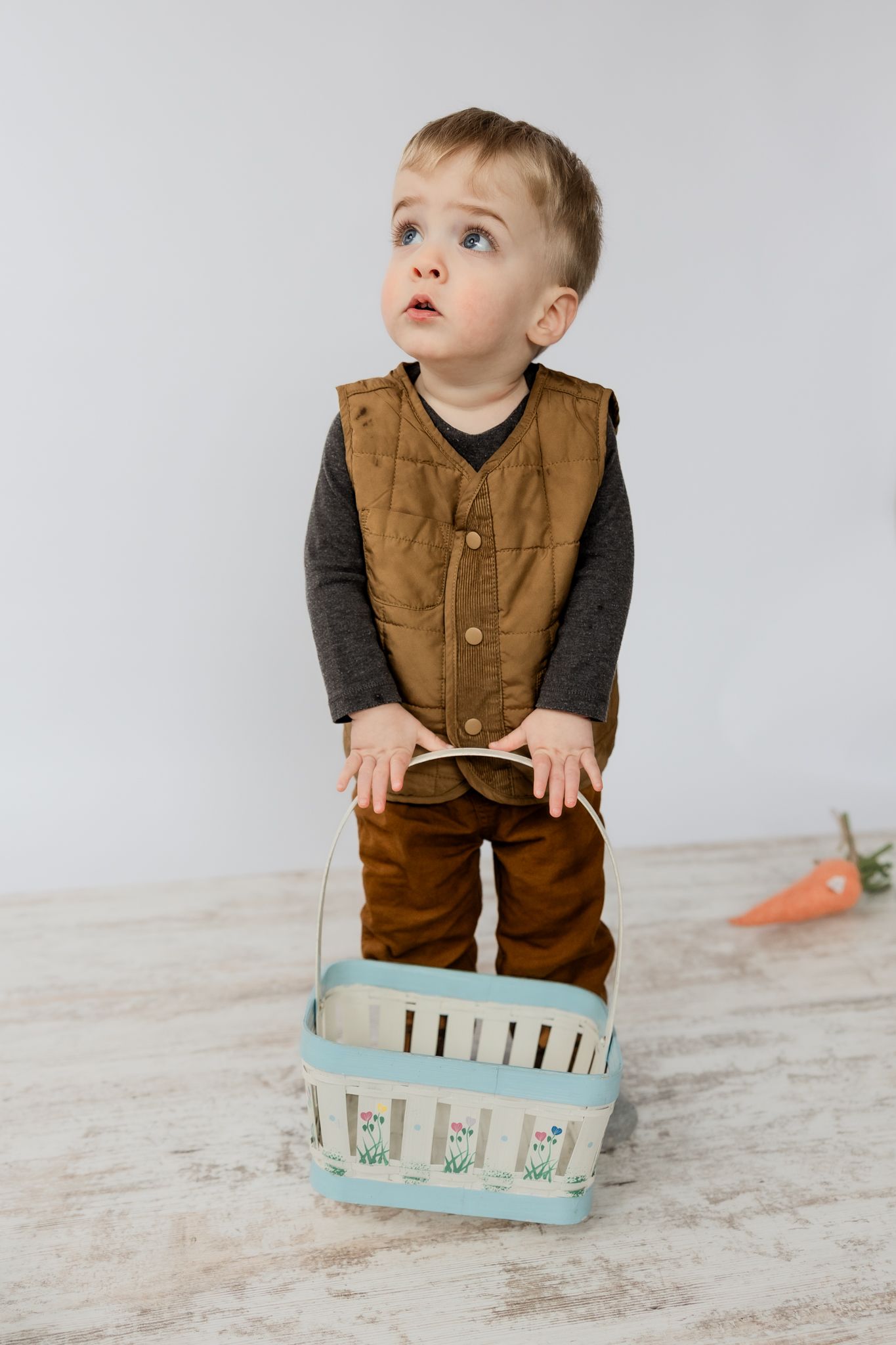 Toddler boy in brown vest and pants holds a decorative Easter basket while looking up curiously.