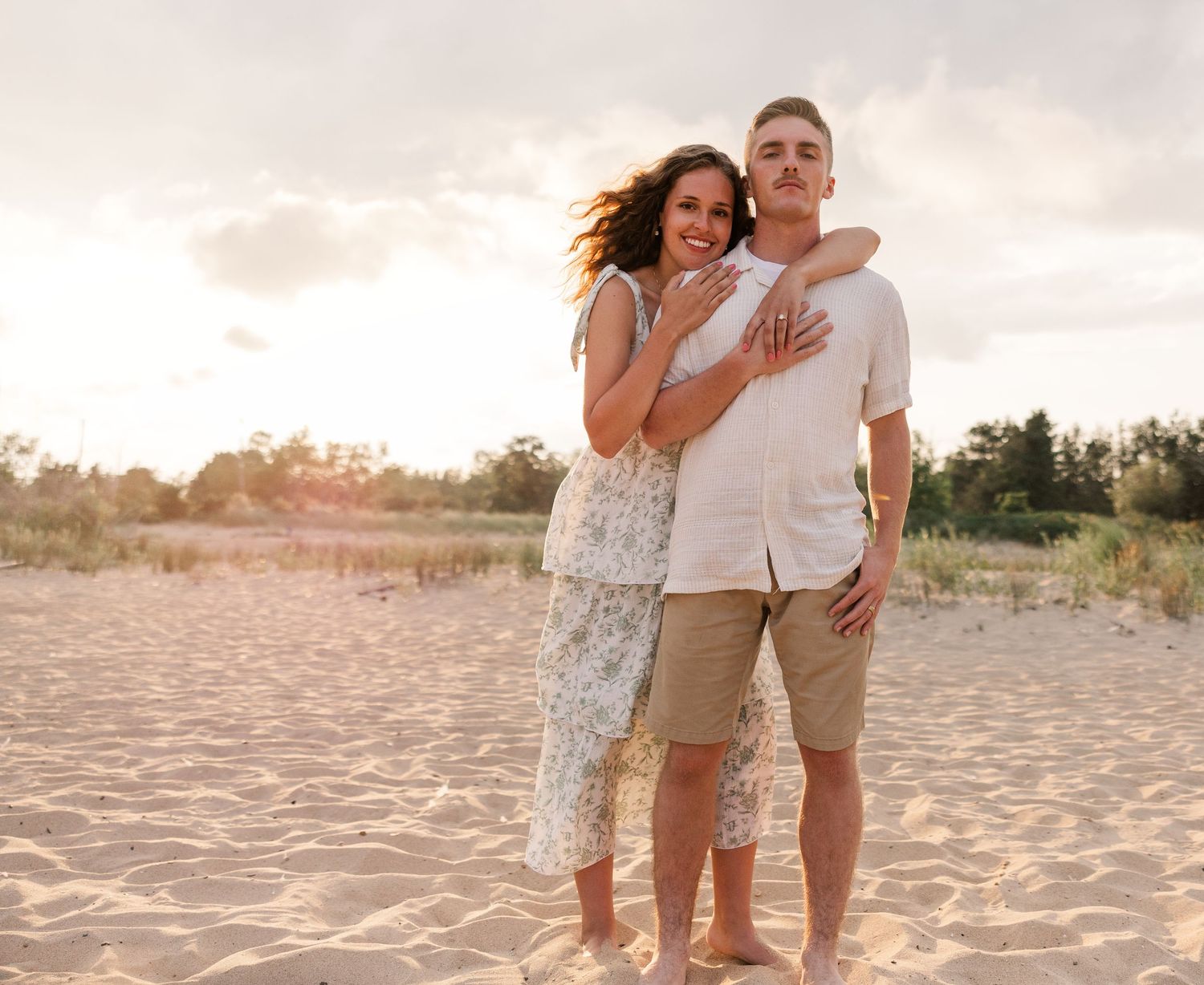 Happy couple embracing on a sandy beach at sunset, woman in floral dress showing engagement ring.