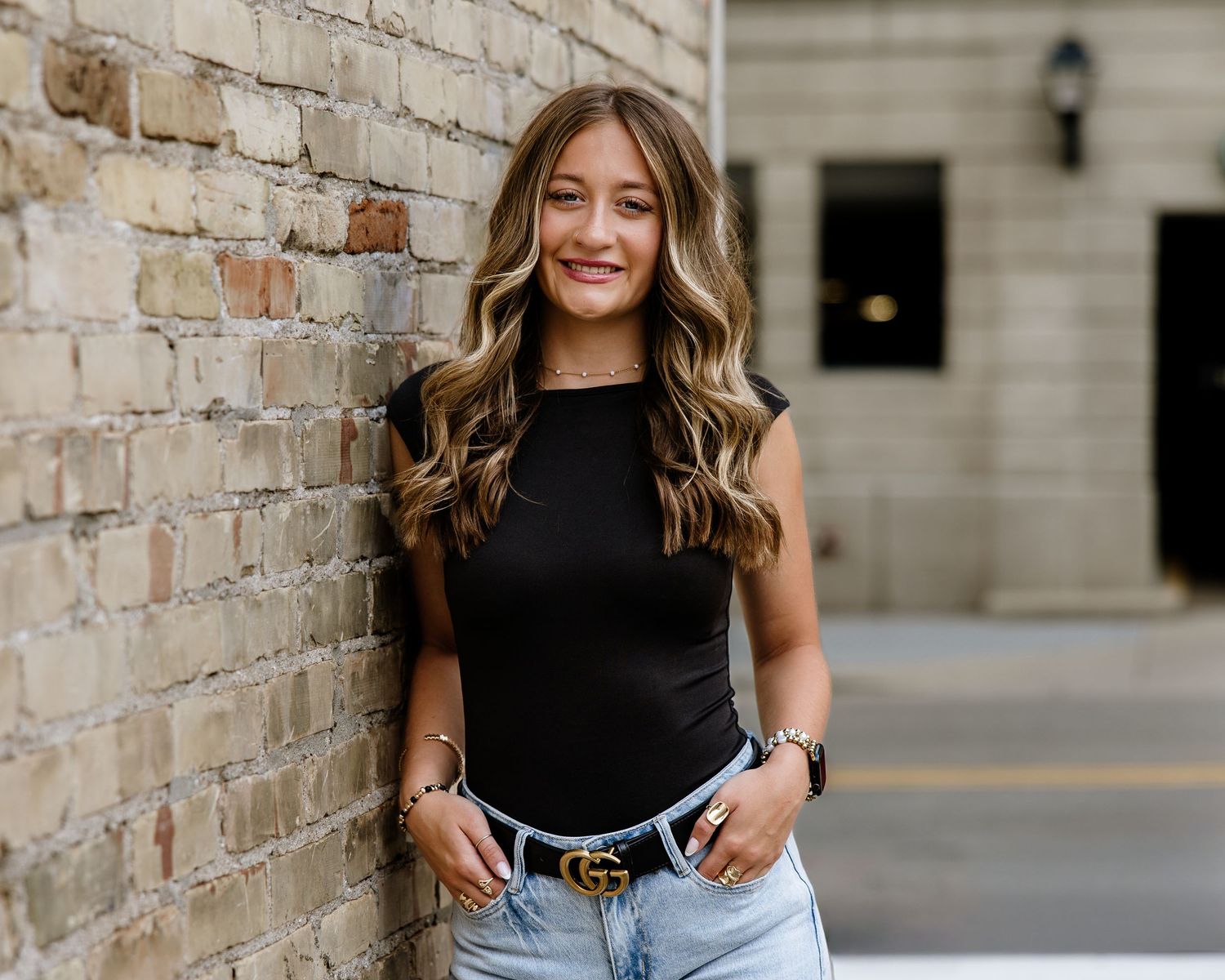 Young woman with wavy blonde hair smiling, wearing black top and light jeans with Gucci belt, leaning against brick wall.