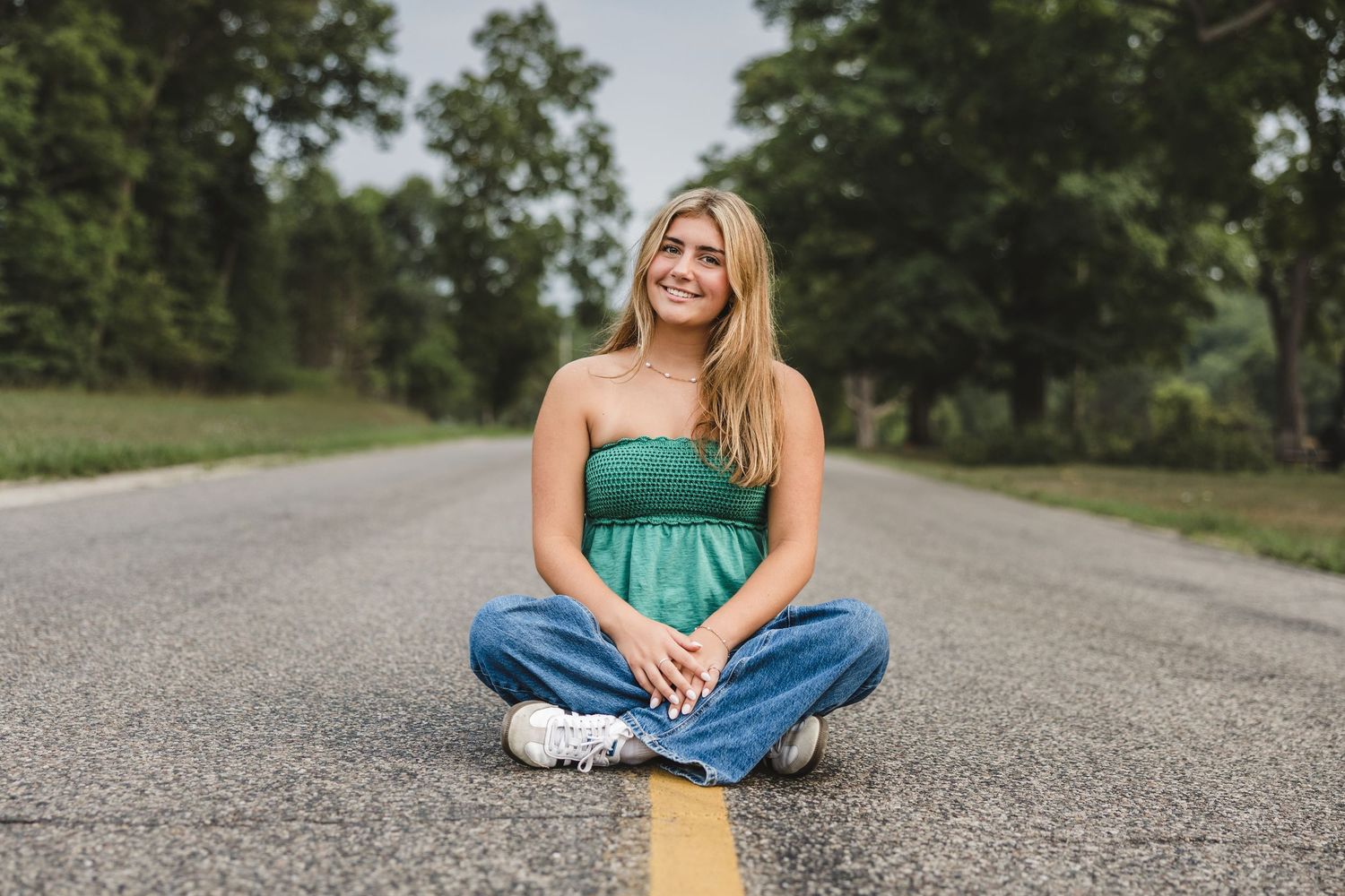 Young woman in green top and jeans sitting cross-legged on a road surrounded by lush green trees, smiling.