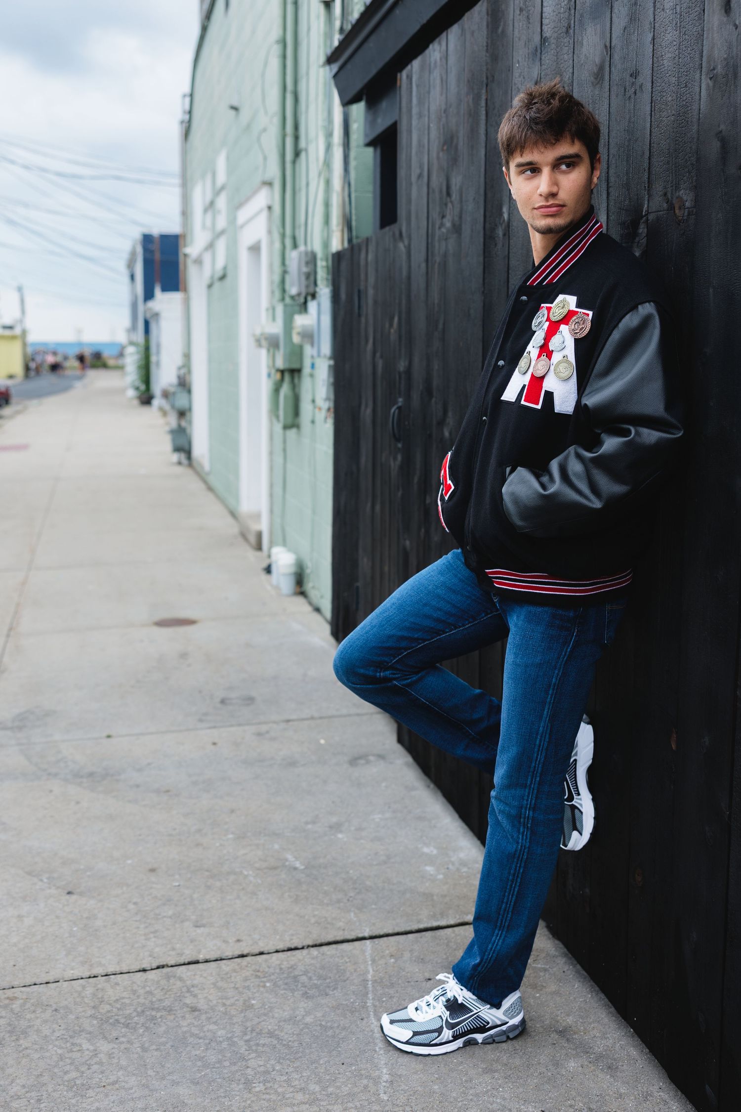 Young man in varsity jacket and jeans leaning against black wall on urban sidewalk.