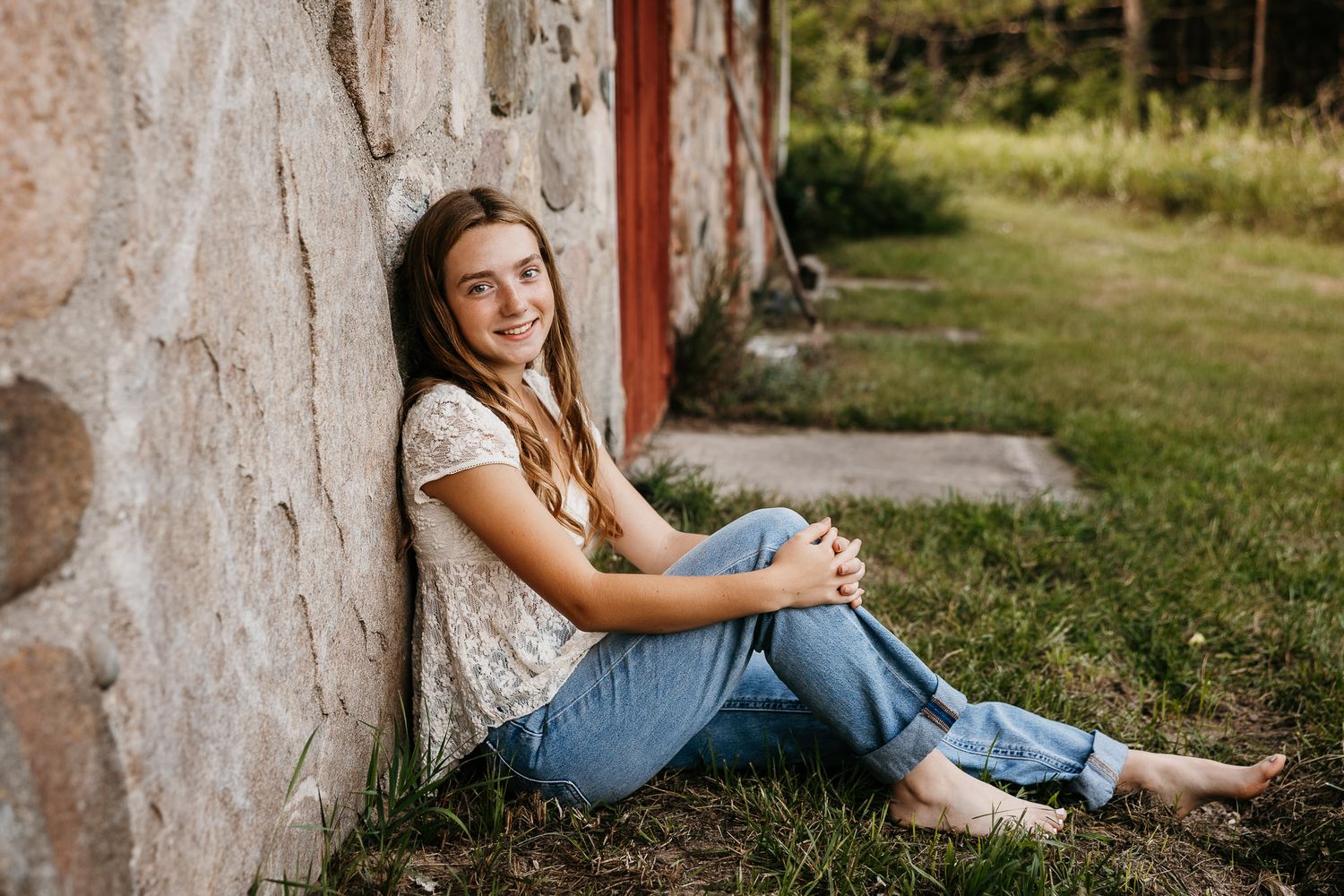 Smiling young woman sitting barefoot on grass, leaning against a stone wall outdoors in jeans and white top.