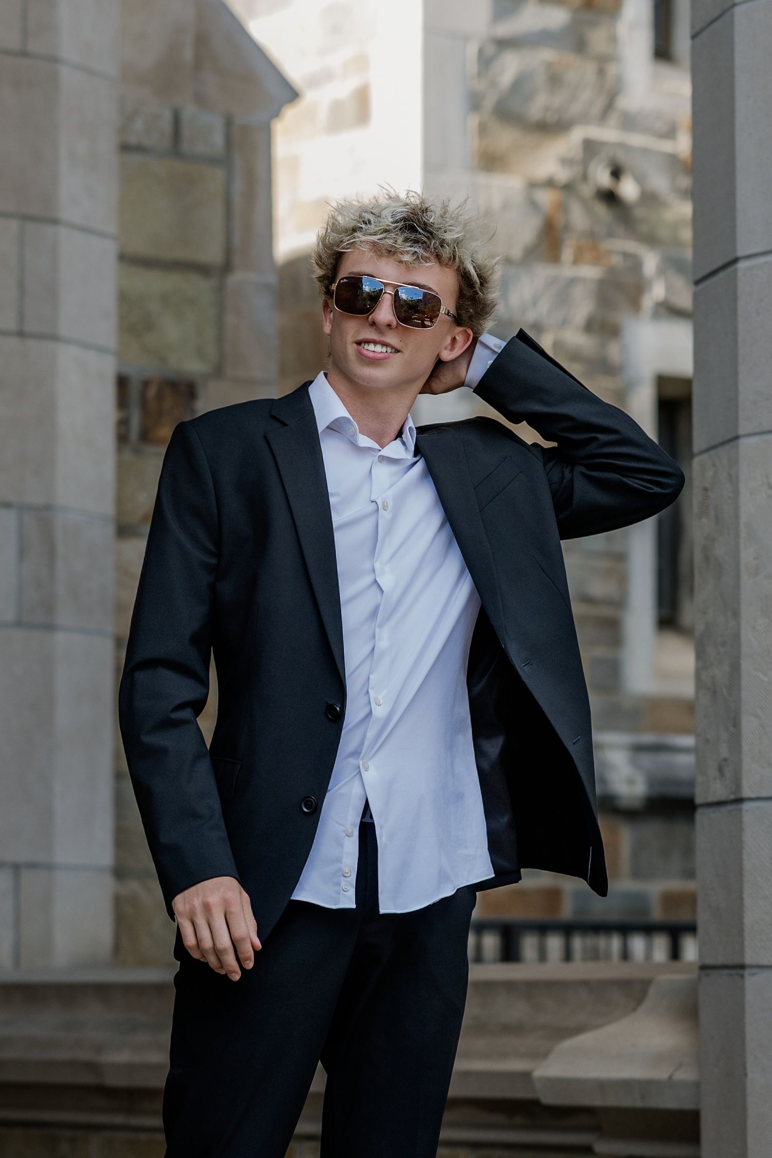 Young man with curly hair wearing sunglasses and a dark suit poses confidently outdoors near stone columns.