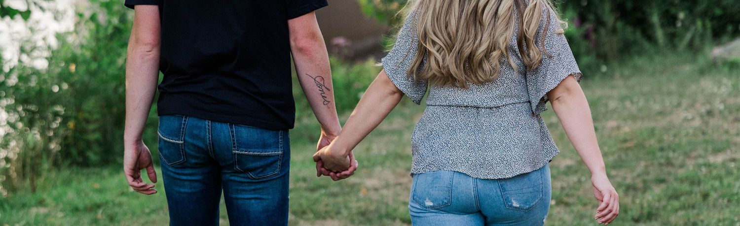 A couple holds hands while walking together outdoors in a green park, viewed from behind wearing casual jeans.