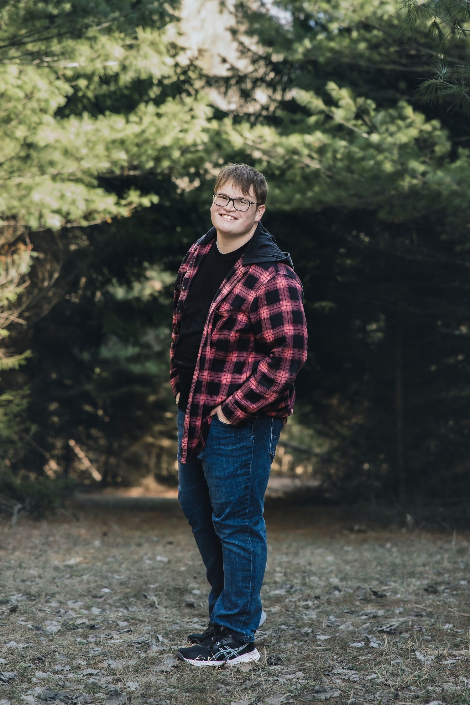Young man in red plaid shirt and jeans smiling outdoors among green pine trees.