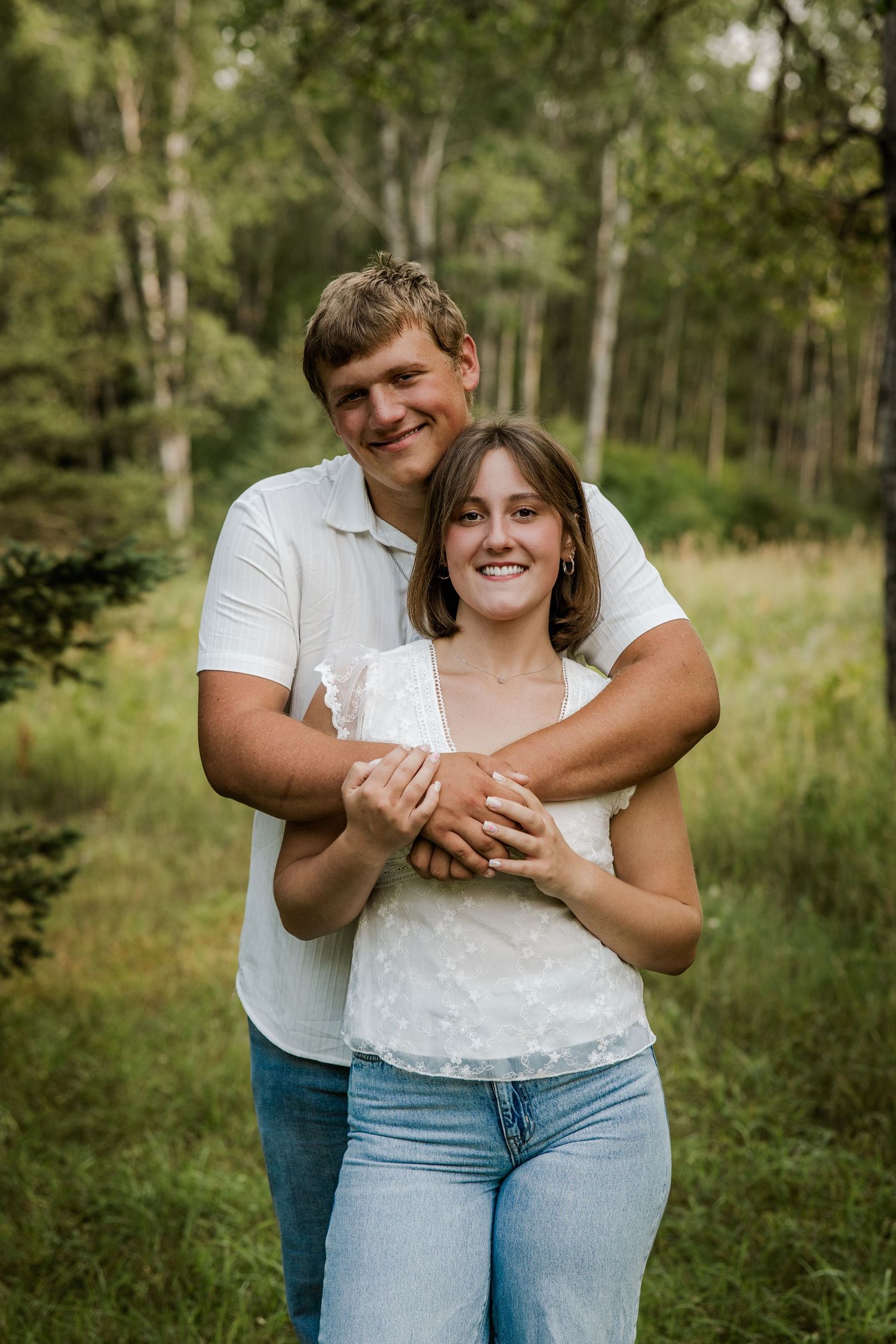 Couple embracing in a lush green forest, wearing white tops and jeans, smiling warmly during an outdoor engagement session.