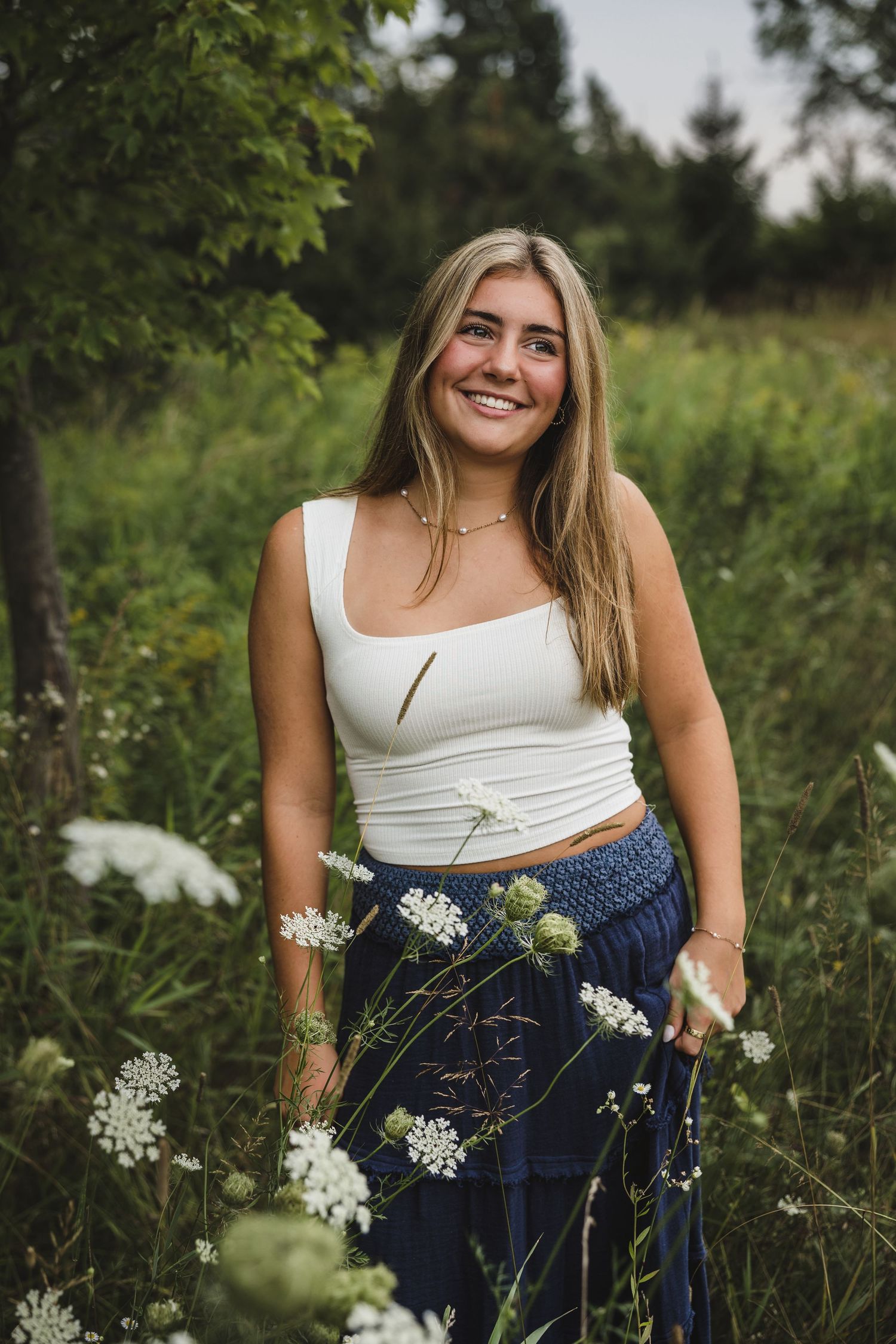 Young blonde woman smiling in a field of white wildflowers, wearing a white crop top and navy blue skirt.