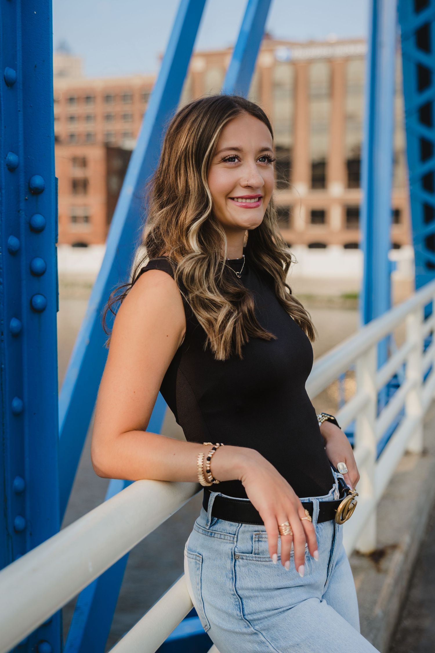 Young woman in black top and jeans smiling on a blue bridge with urban buildings in the background.