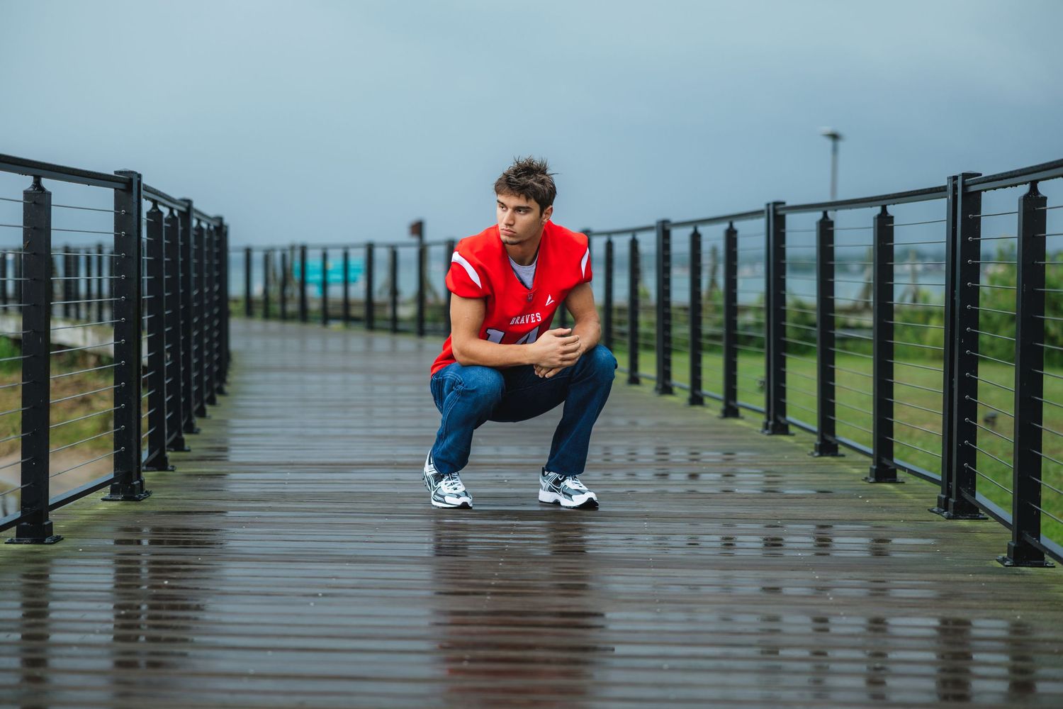 Young man in red jersey squatting on wet boardwalk during overcast day, wearing jeans and sneakers.