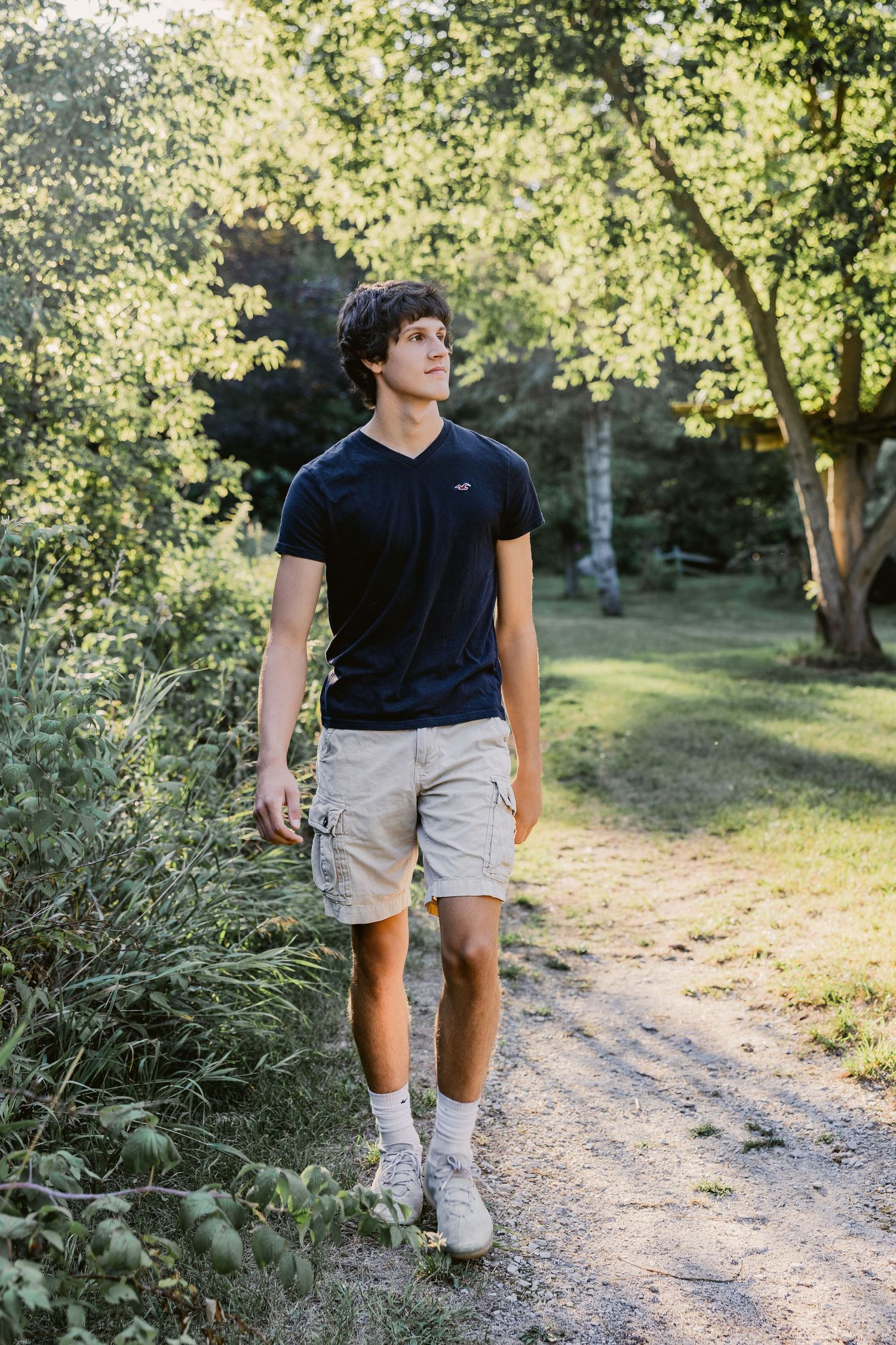 Young man in navy t-shirt and khaki cargo shorts walking on a sunlit forest path surrounded by lush green trees.
