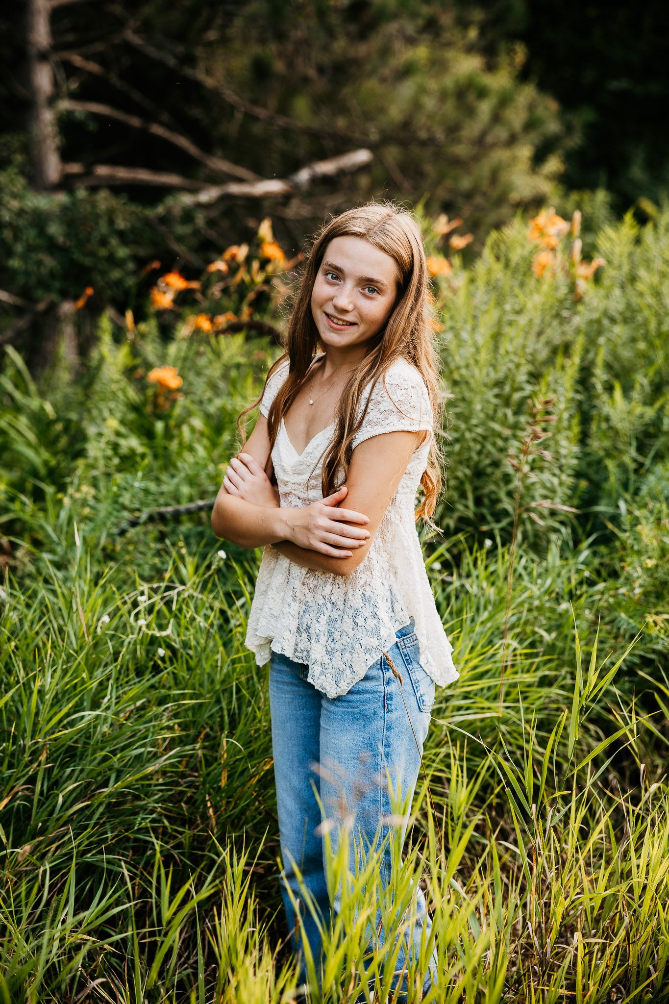Young woman standing in lush green garden with orange lilies, wearing lace top and jeans, arms crossed.
