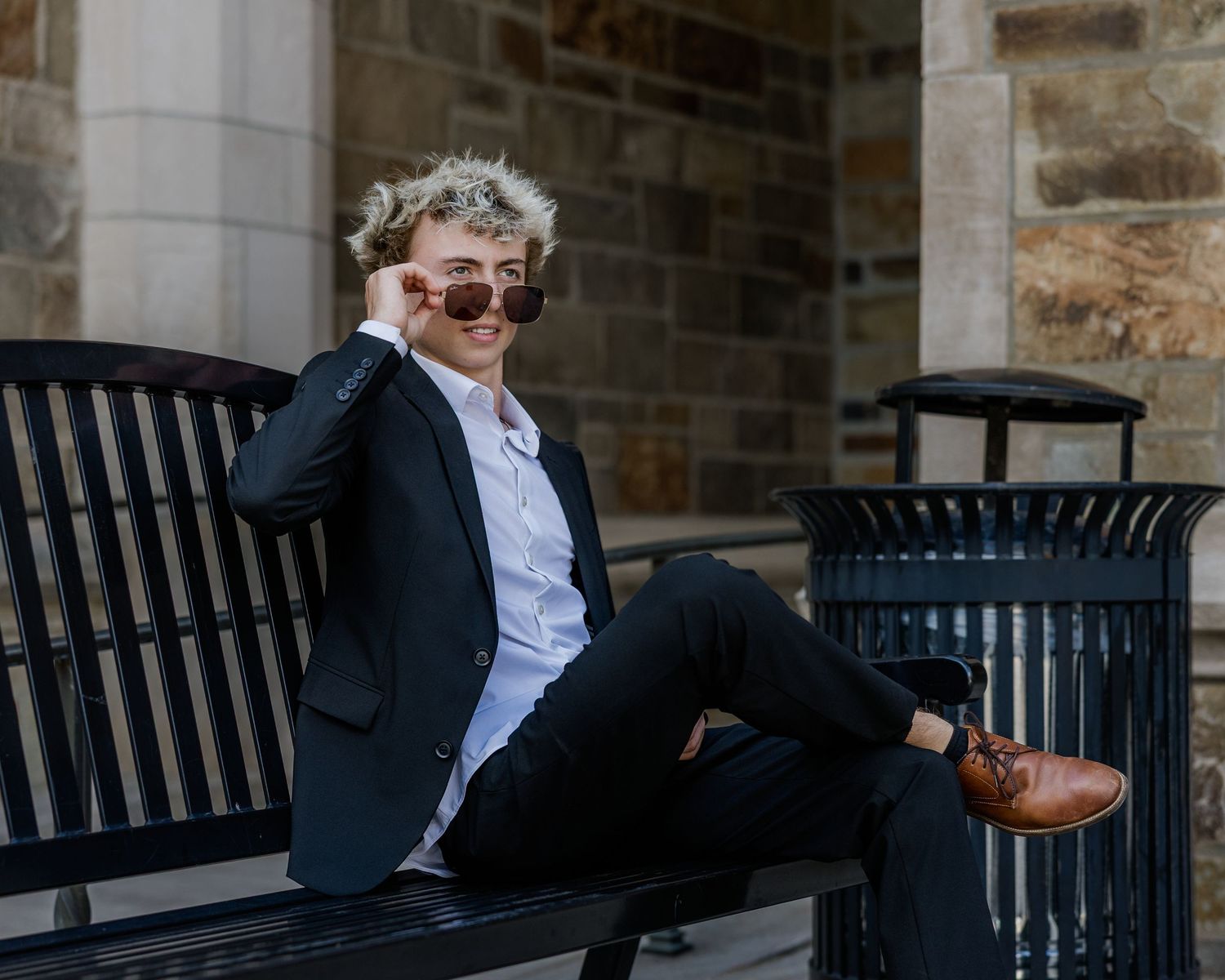 Young man in black blazer and sunglasses relaxing on a bench outdoors near stone architecture.