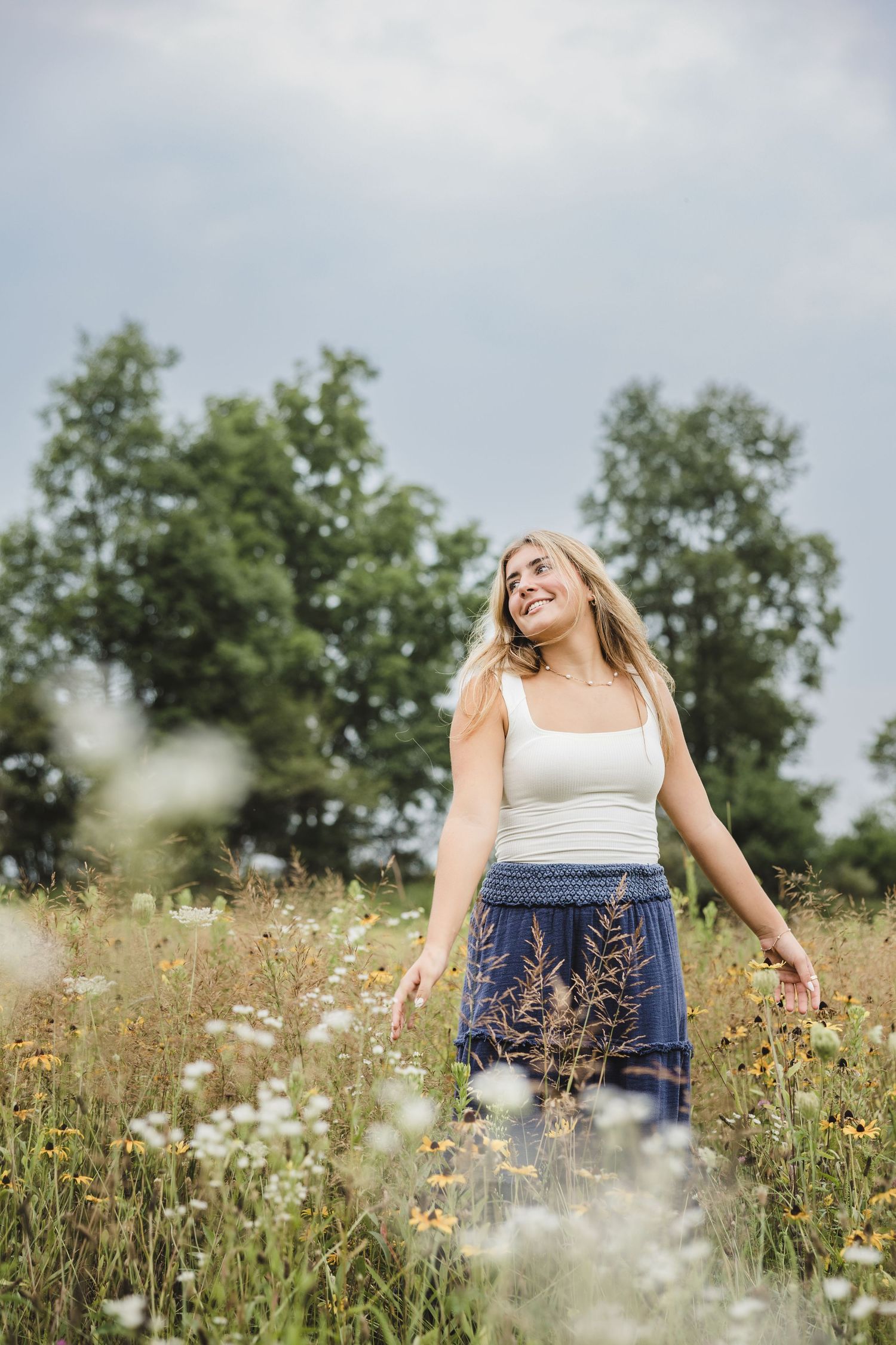 Smiling blonde woman in white tank top and blue floral skirt standing in a wildflower meadow on a cloudy day.