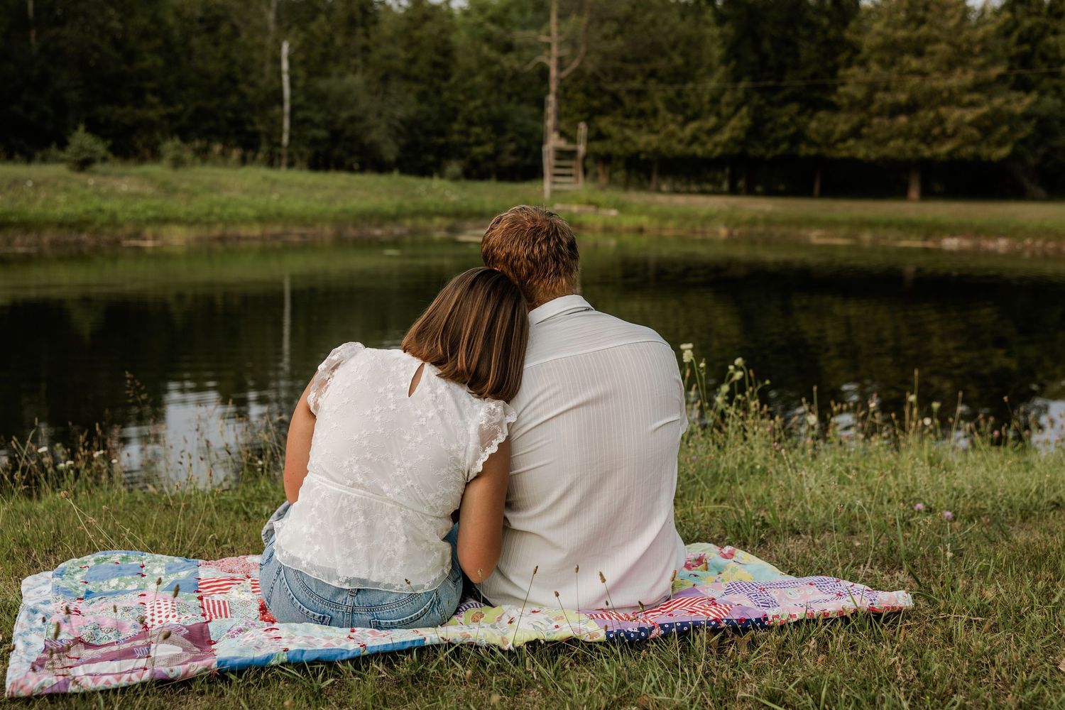 Couple sitting together on a colorful quilt by a serene pond surrounded by lush green trees.