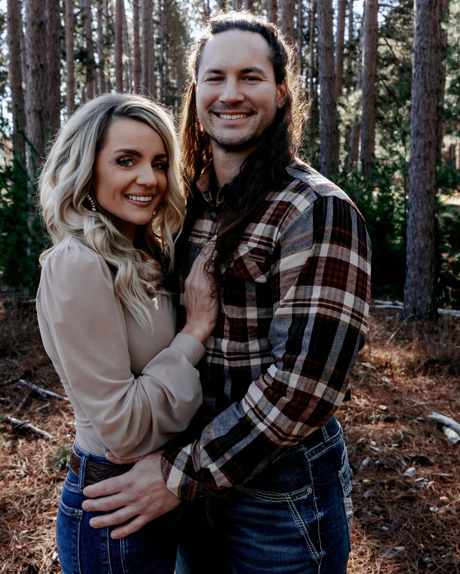 A smiling couple poses together in a pine forest, woman in beige top and man in plaid flannel shirt with long hair.