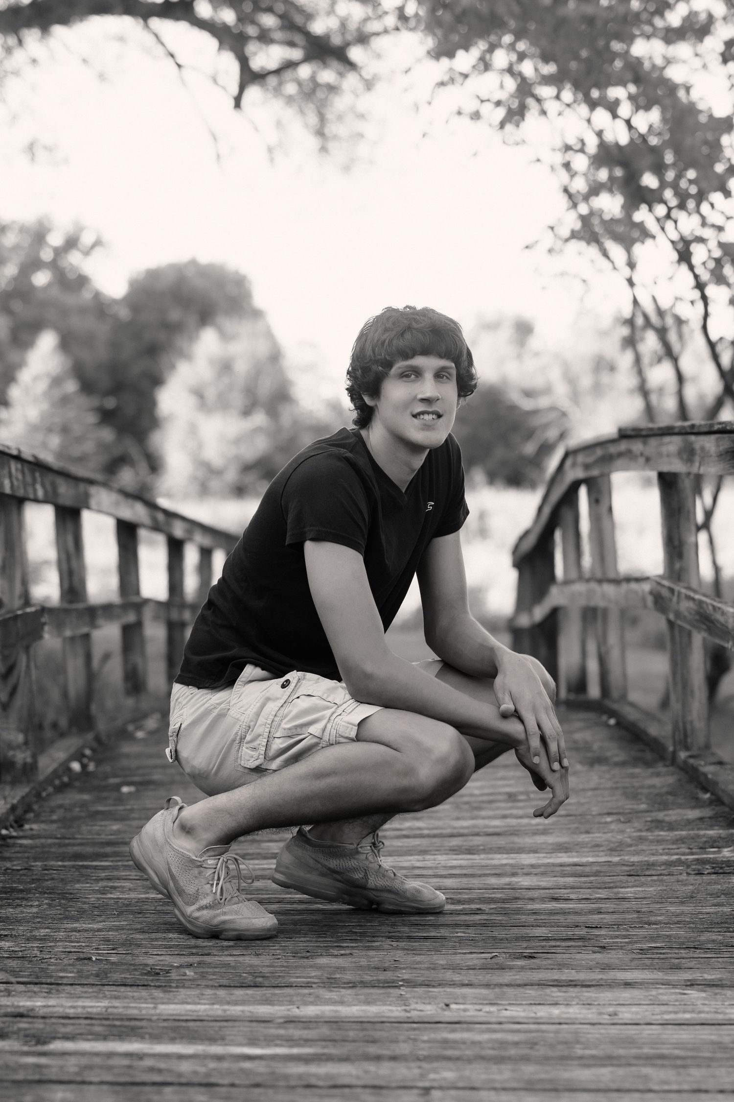 Black and white photo of a young man crouching on a wooden bridge, smiling, wearing shorts and a black t-shirt.