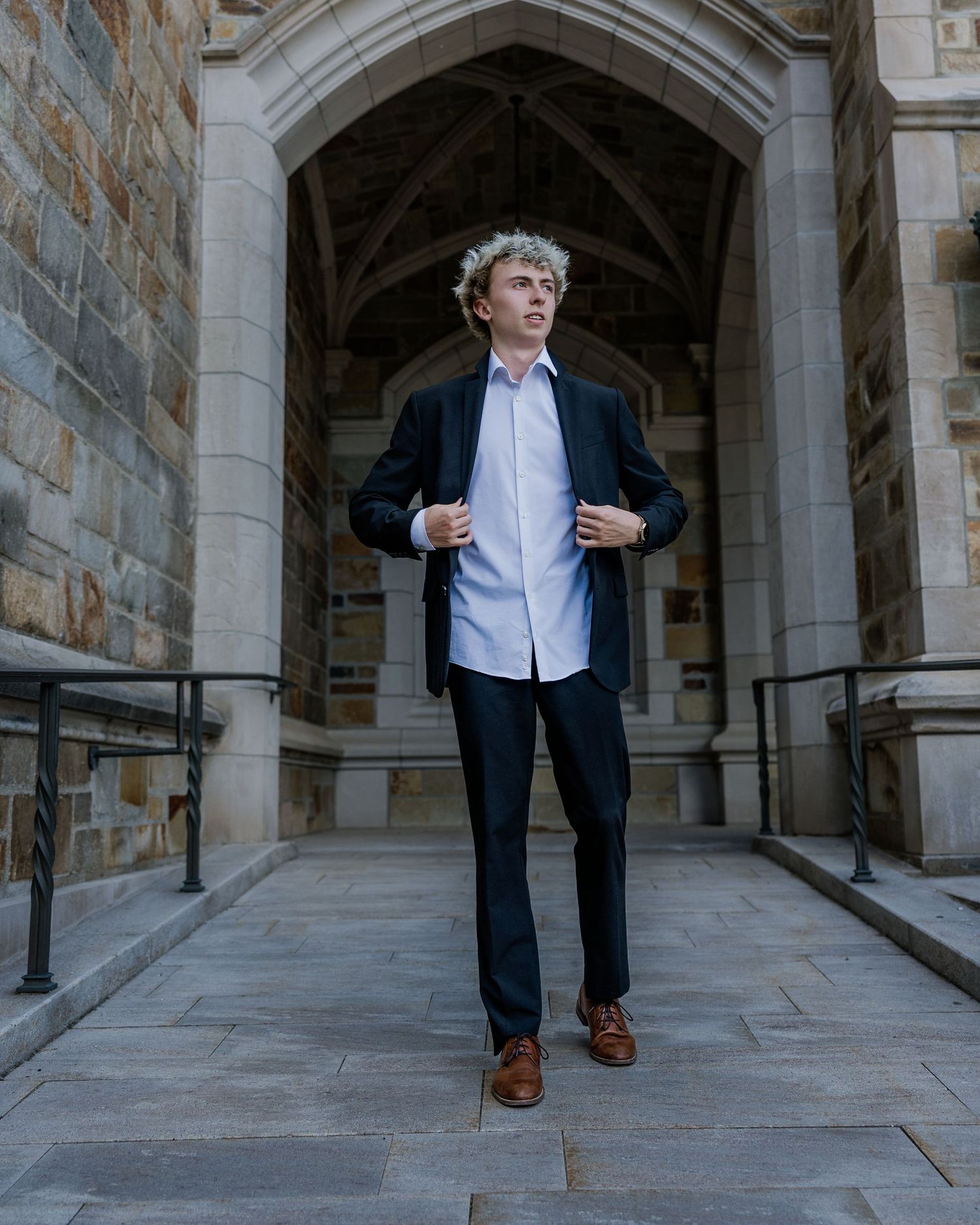 Young man in dark suit and light blue shirt stands confidently under stone archway of classic university building.