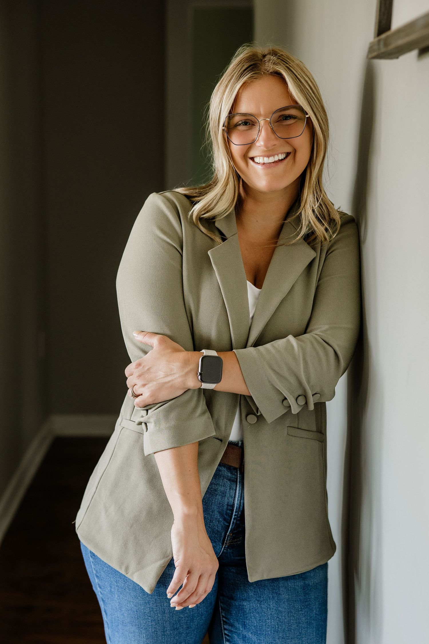 Smiling blonde woman in olive blazer and jeans leans against wall wearing sunglasses and smartwatch.
