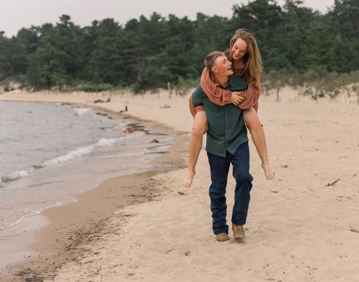 Couple enjoying a piggyback ride on a sandy beach with trees in the background.