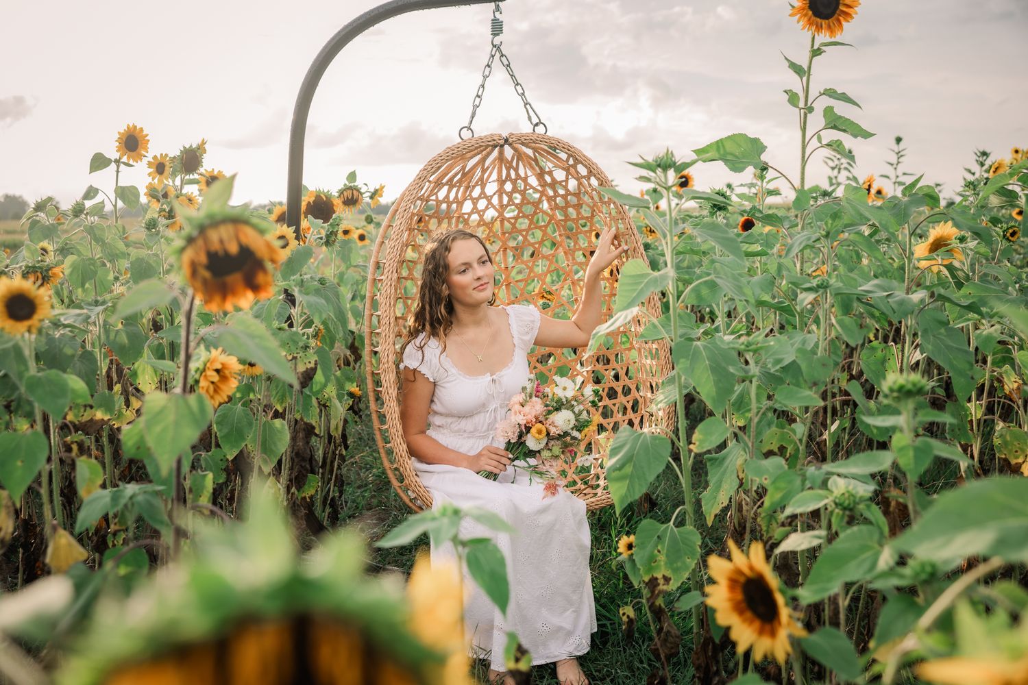 Woman in white dress sitting in wicker swing chair surrounded by blooming sunflower field holding a bouquet.