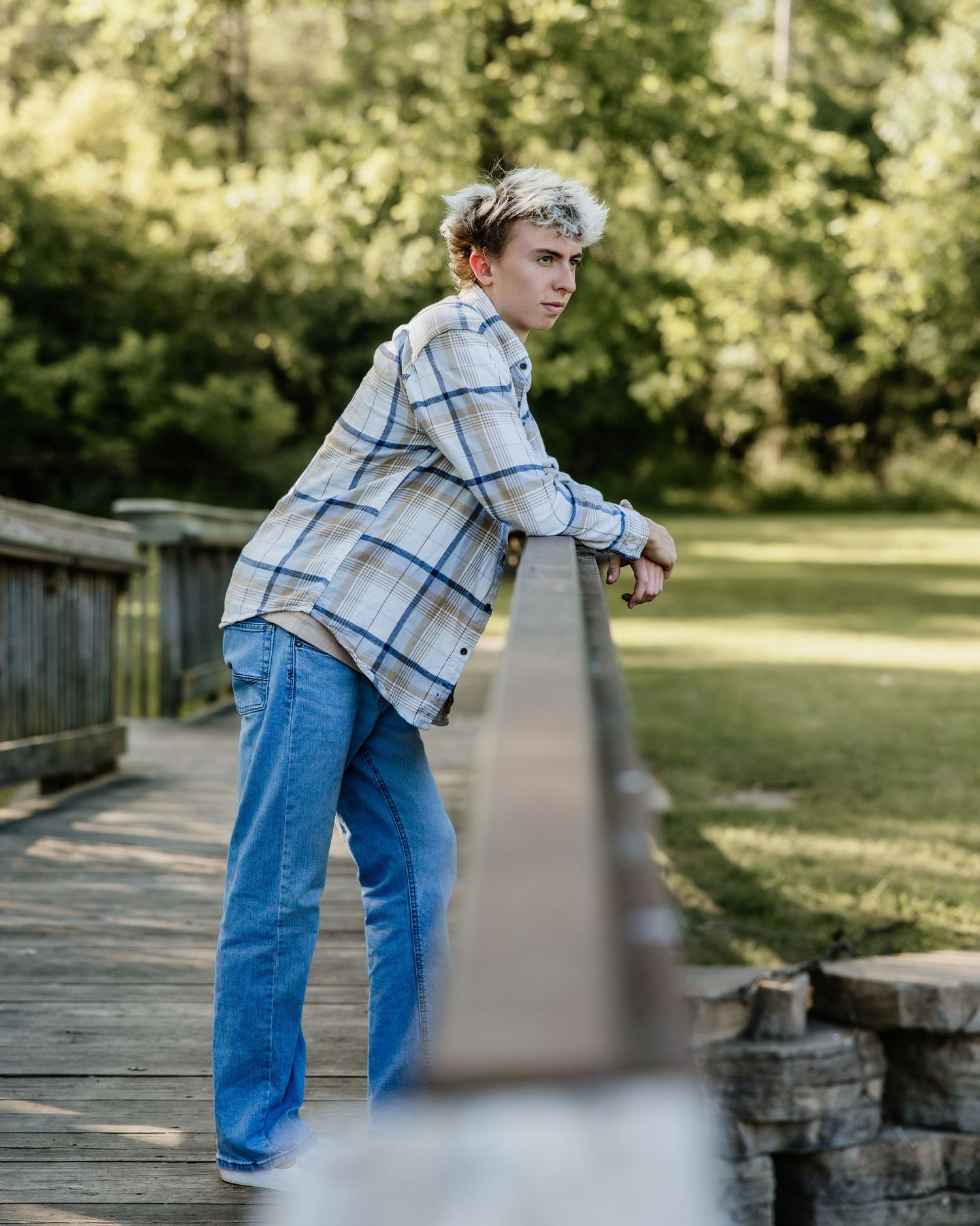 Young man in plaid shirt and jeans leaning on wooden bridge railing in a lush green park setting.