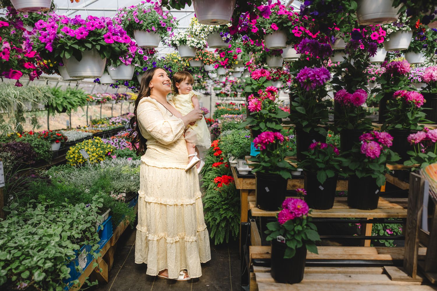 Woman in cream dress holding baby among vibrant pink flowers and hanging baskets in a lush greenhouse.
