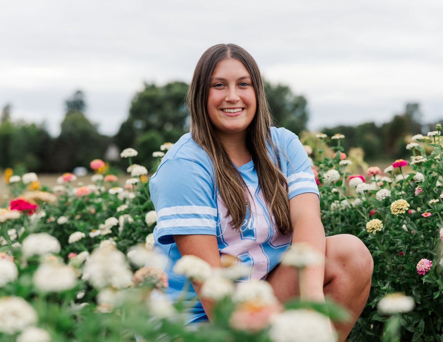 Smiling brunette teen girl crouches among colorful zinnia flowers in a garden under cloudy skies.