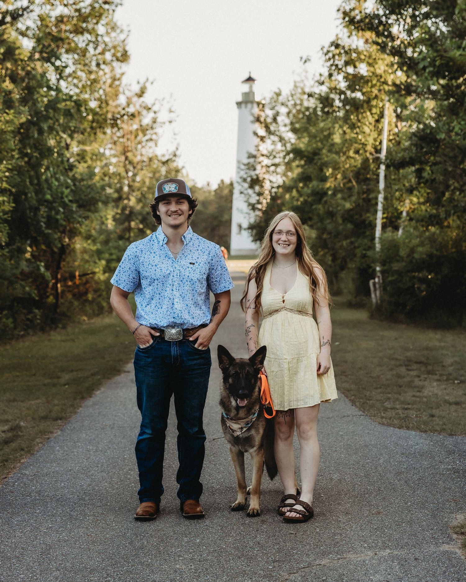 A young couple stands smiling on a tree-lined path with their German Shepherd dog on an orange leash near a lighthouse.