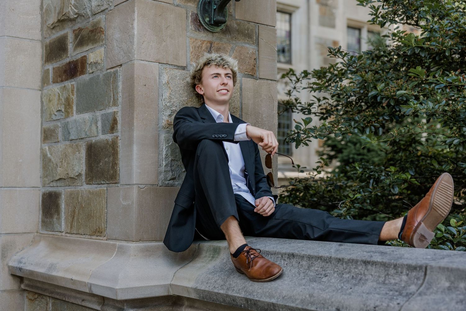 Young man in suit sitting relaxed on stone ledge outside historic building, looking up thoughtfully.