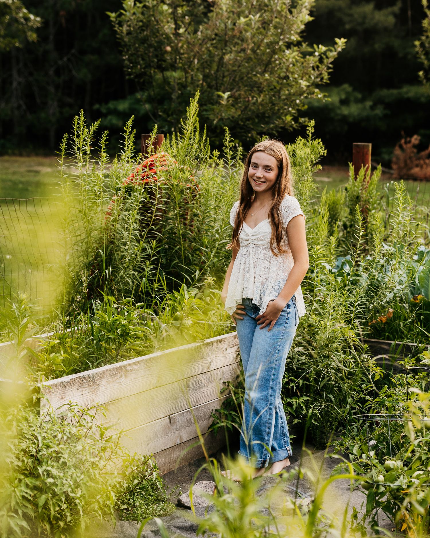 Young woman in white top and jeans smiles in a lush garden with raised wooden planter beds and wildflowers.