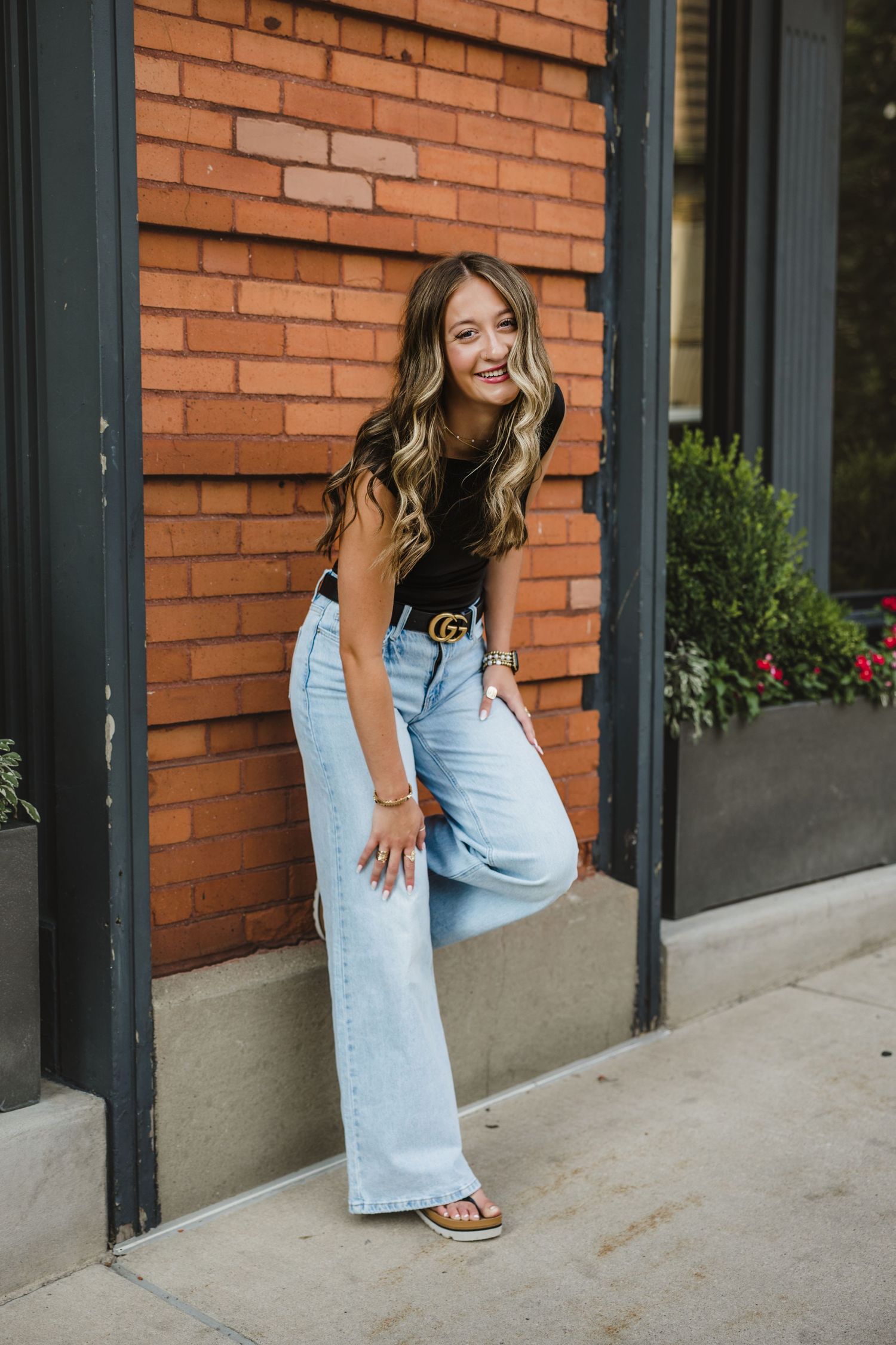 Young woman in wide-leg jeans and black top with Gucci belt poses against a red brick wall outdoors.