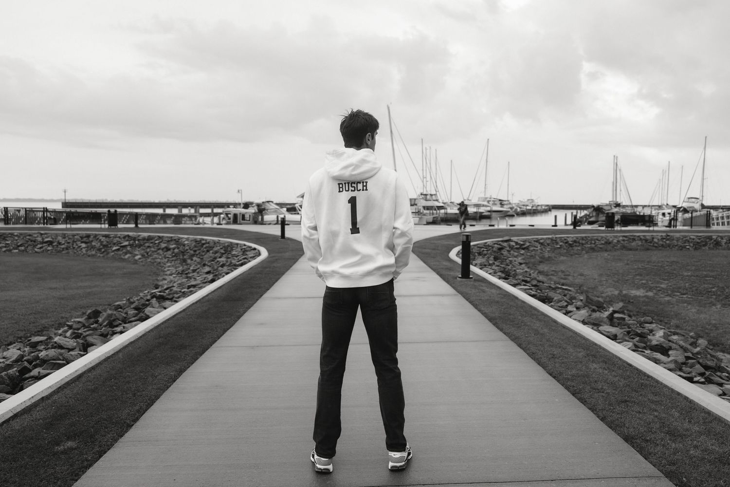 Black and white photo of a person wearing a jersey numbered 1 standing on a marina walkway with boats in the background.