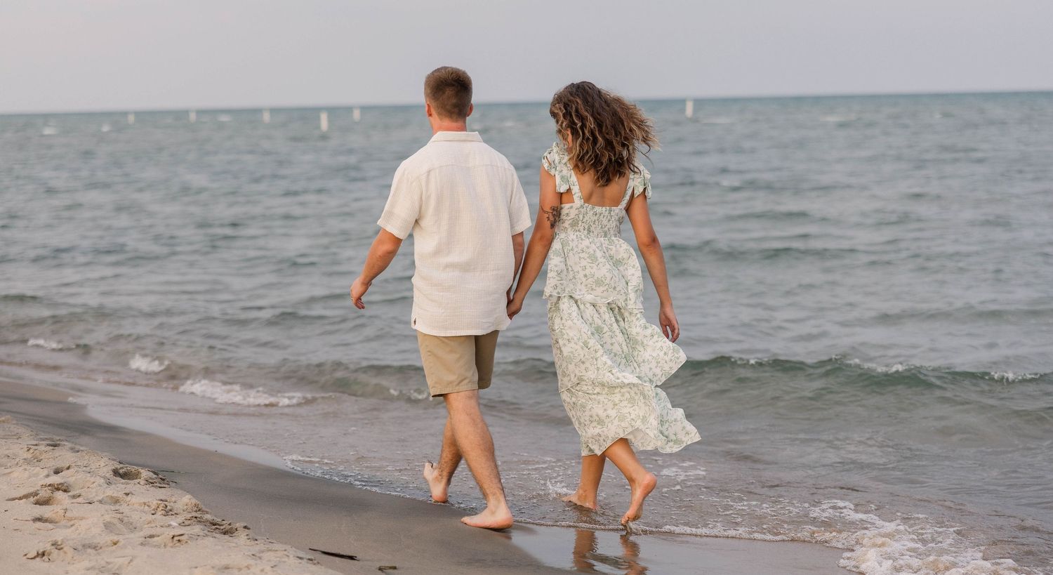 Couple walking hand in hand along a sandy beach shoreline, wearing casual summer attire on a calm, cloudy day.