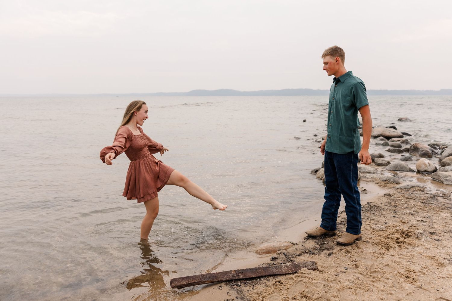 Couple laughing on rocky shoreline, woman in rust dress splashing water, man in teal shirt smiling, misty lake backdrop.