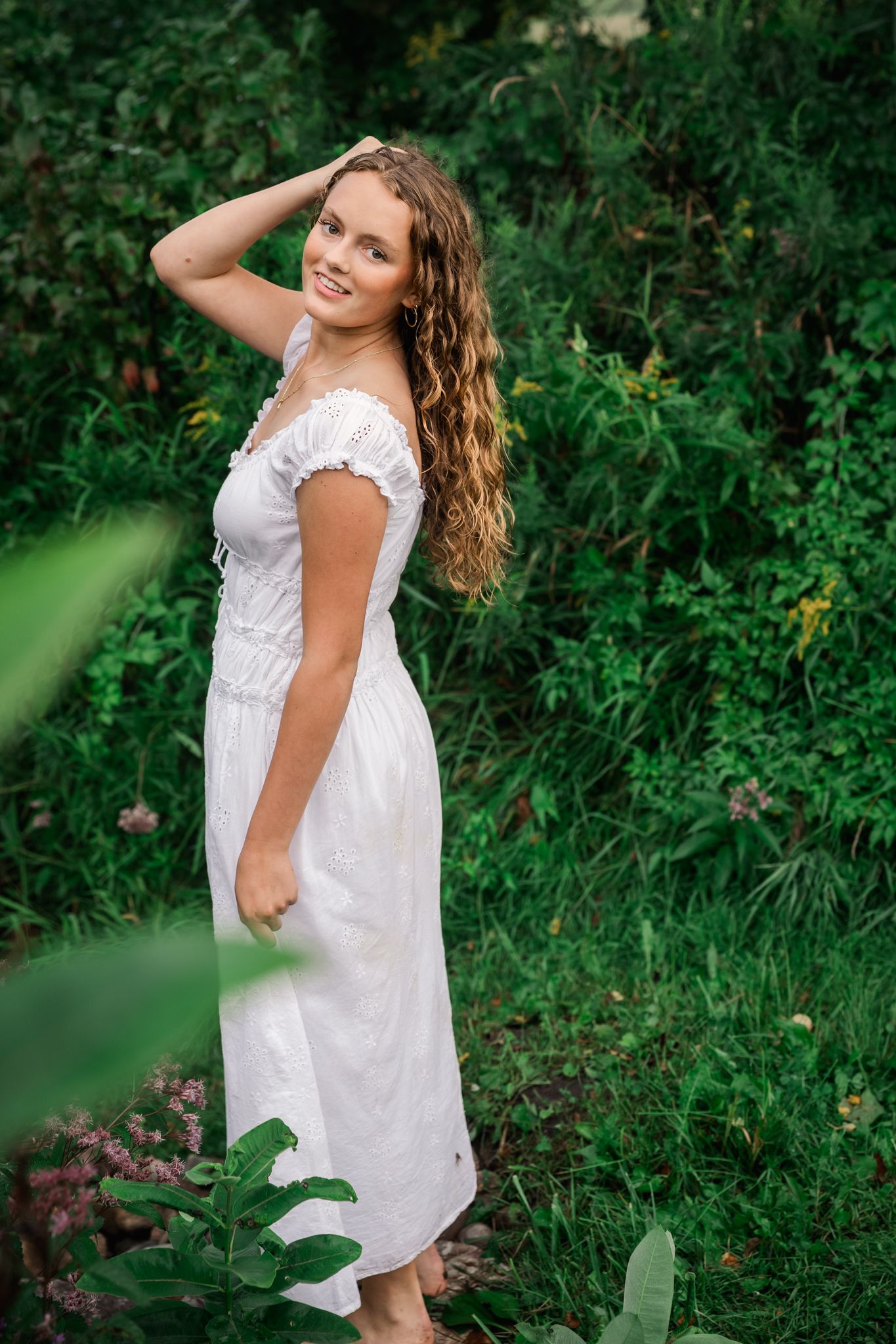 Young woman in white off-shoulder dress smiling outdoors among lush green foliage and plants.