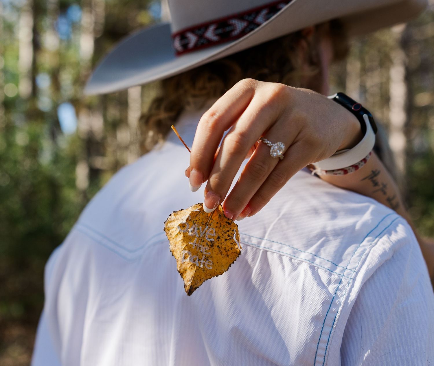 Close-up of woman holding autumn leaf showing diamond engagement ring and tattoos outdoors.