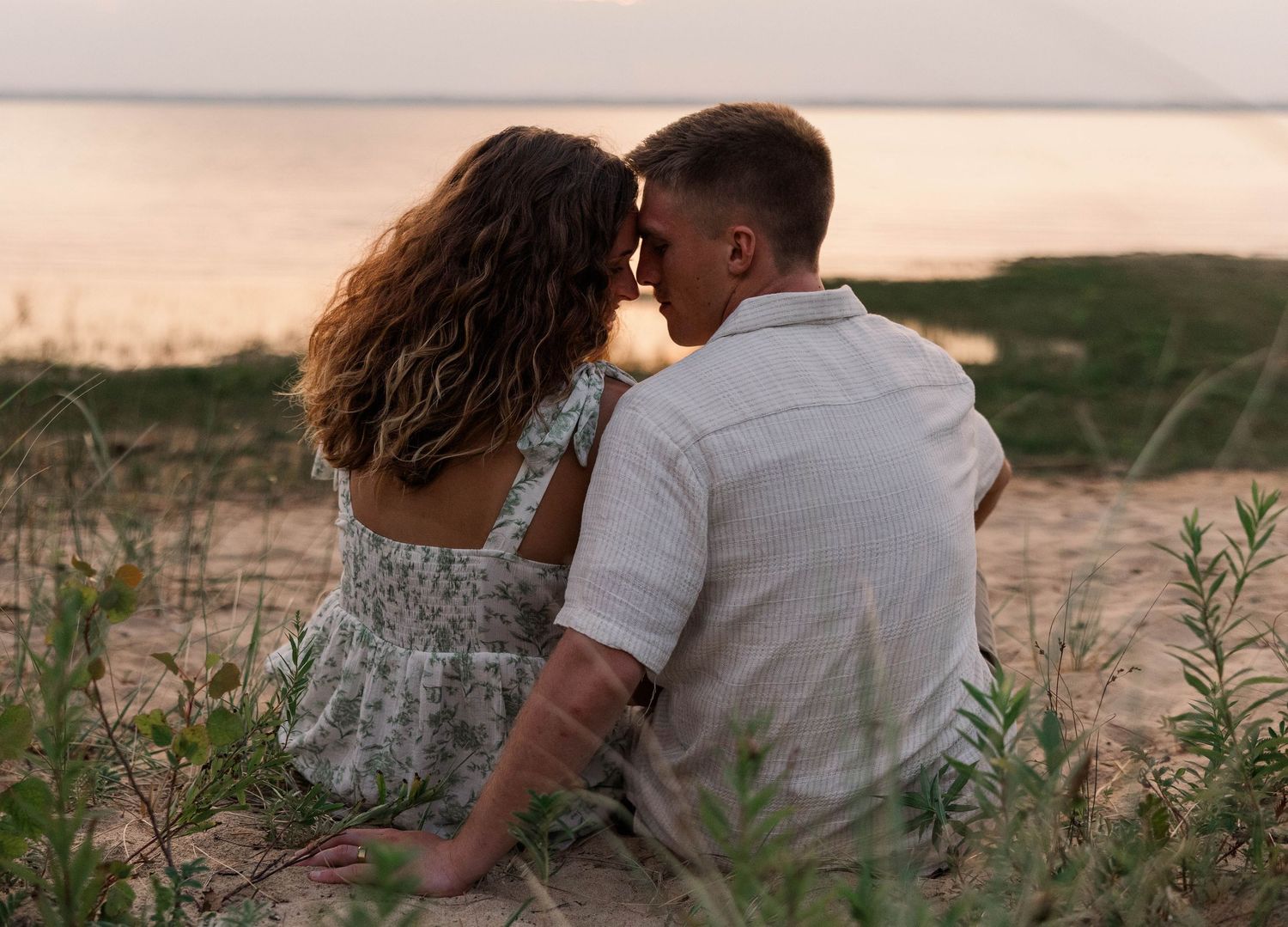 Couple sits together on sandy beach at sunset, touching foreheads in a romantic embrace.