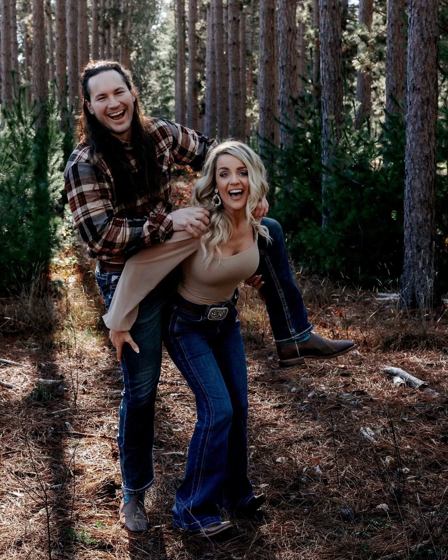 Laughing couple in pine forest, man lifting woman in jeans and cowboy boots playfully.